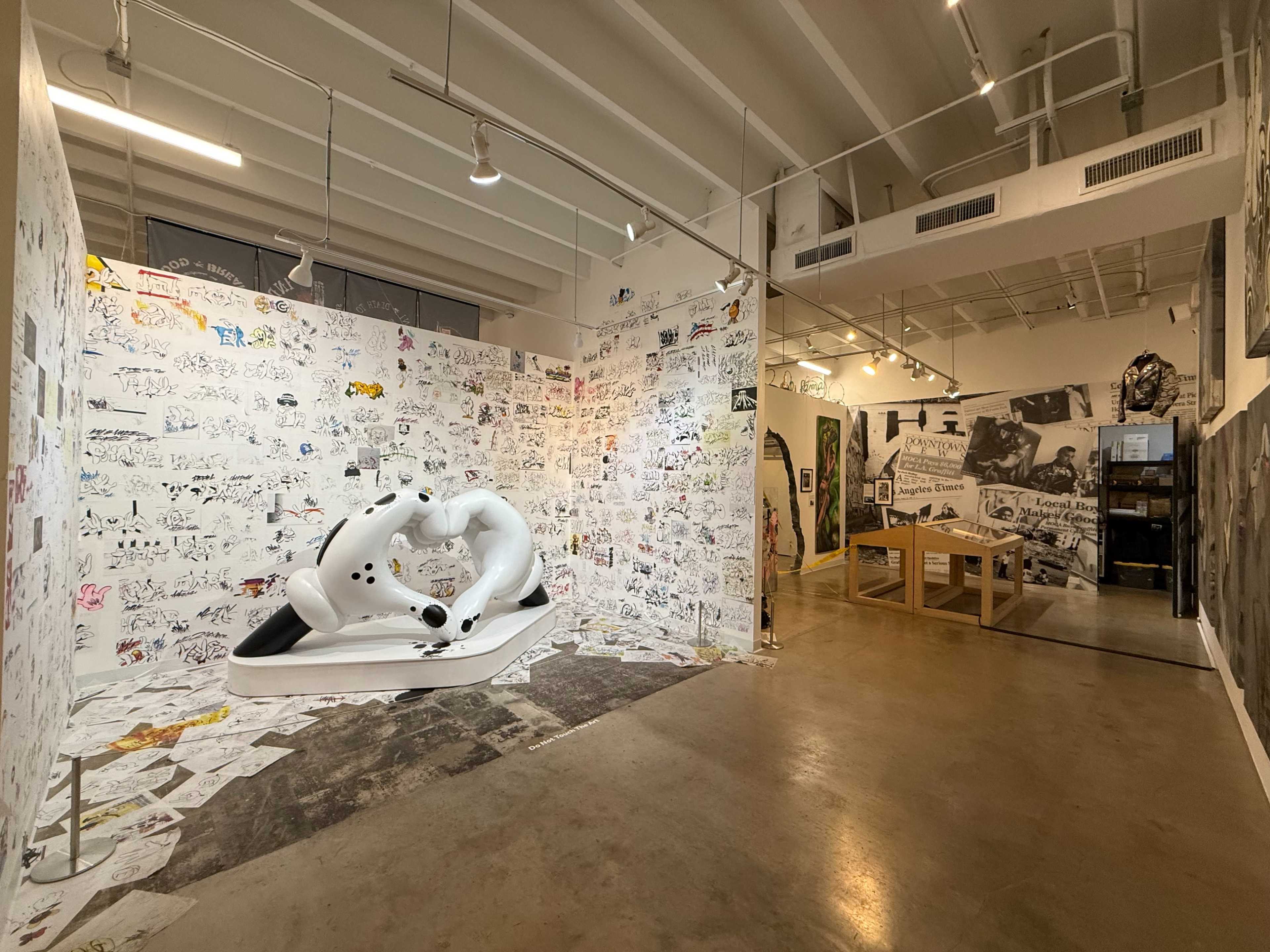 The image shows an art gallery featuring a white sculpture on a raised platform surrounded by walls covered in drawings and sketches, with a wooden table positioned in the background.
