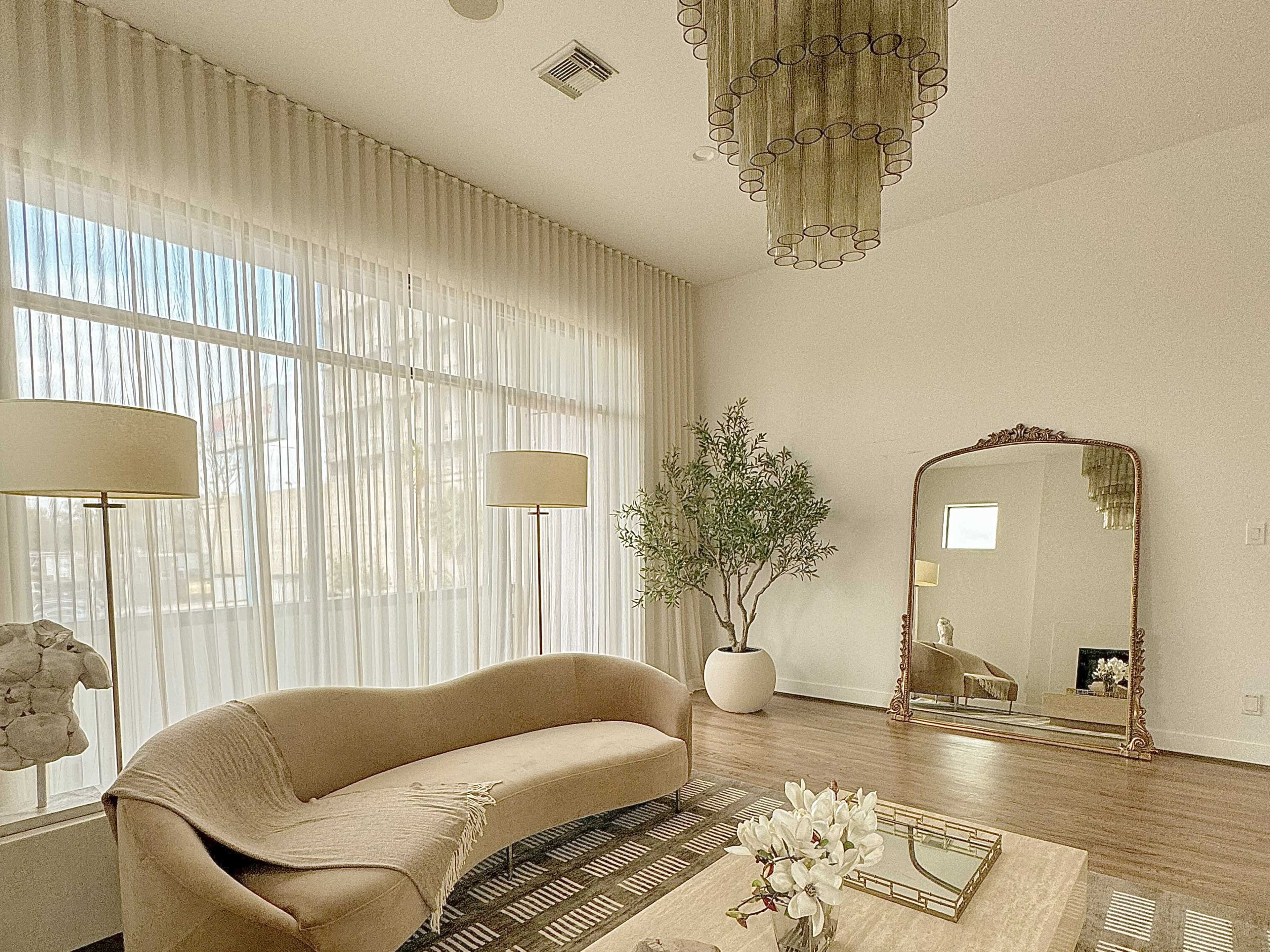 The image shows a modern living space featuring a curved beige sofa, a large mirror, a potted plant, two lamps, and a decorative coffee table with flowers, all illuminated by natural light through sheer curtains.