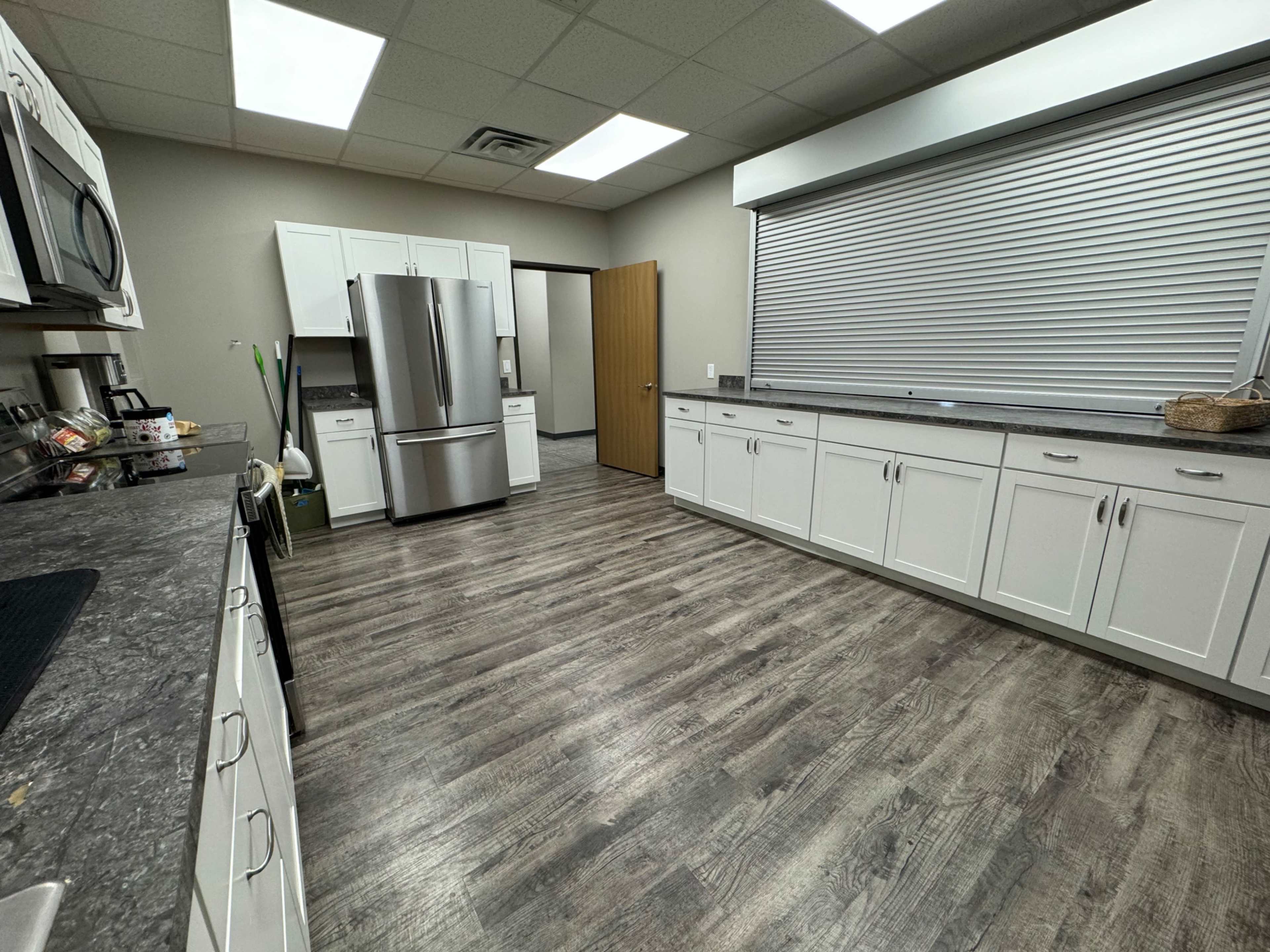 A modern kitchen features white cabinets, stainless steel appliances, and gray laminate flooring under a window with a roll-up shade.