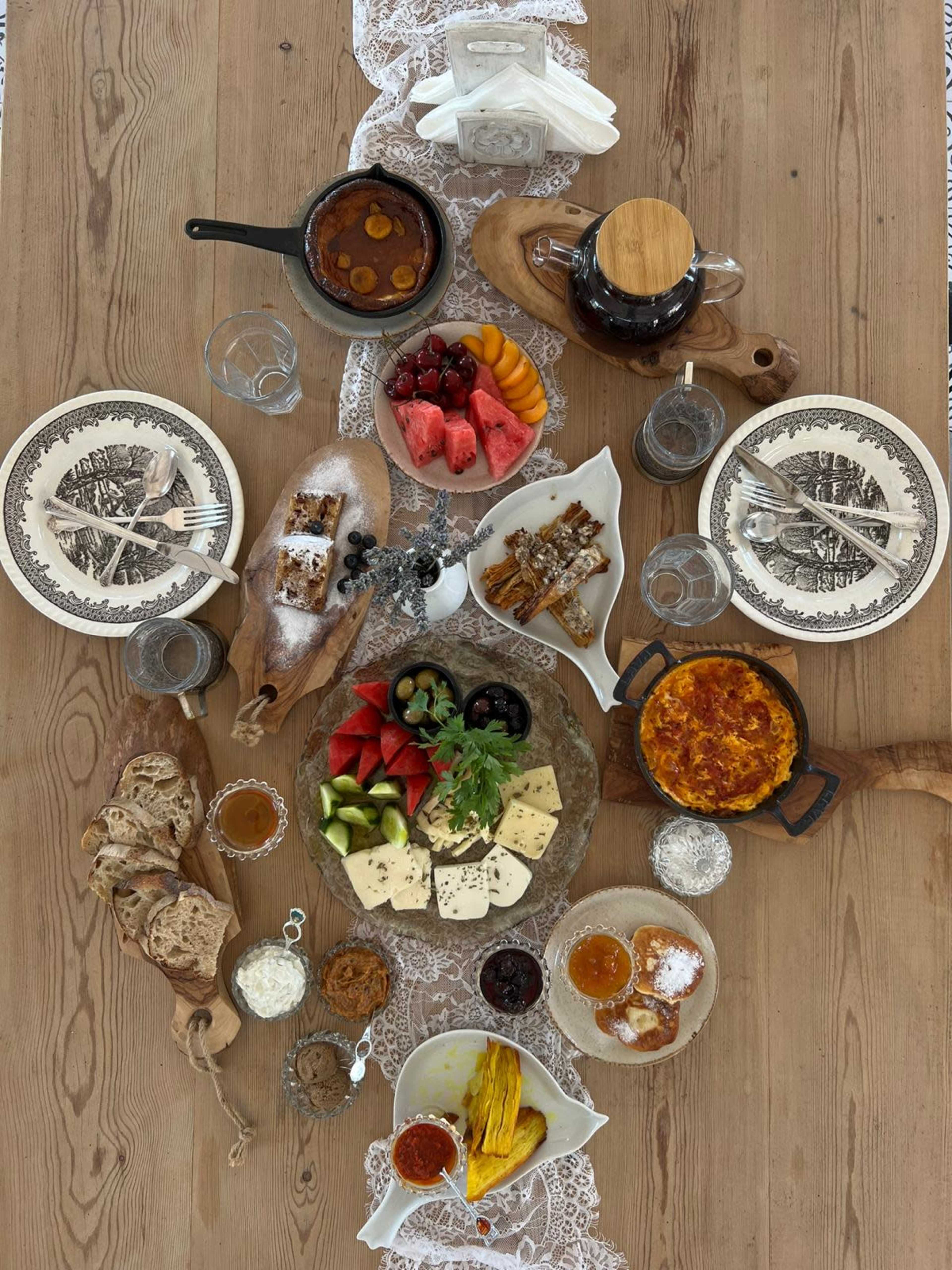 A large wooden table is set with an assortment of breakfast foods including cheeses, fruits, breads, and pastries, accompanied by cutlery and drinks.