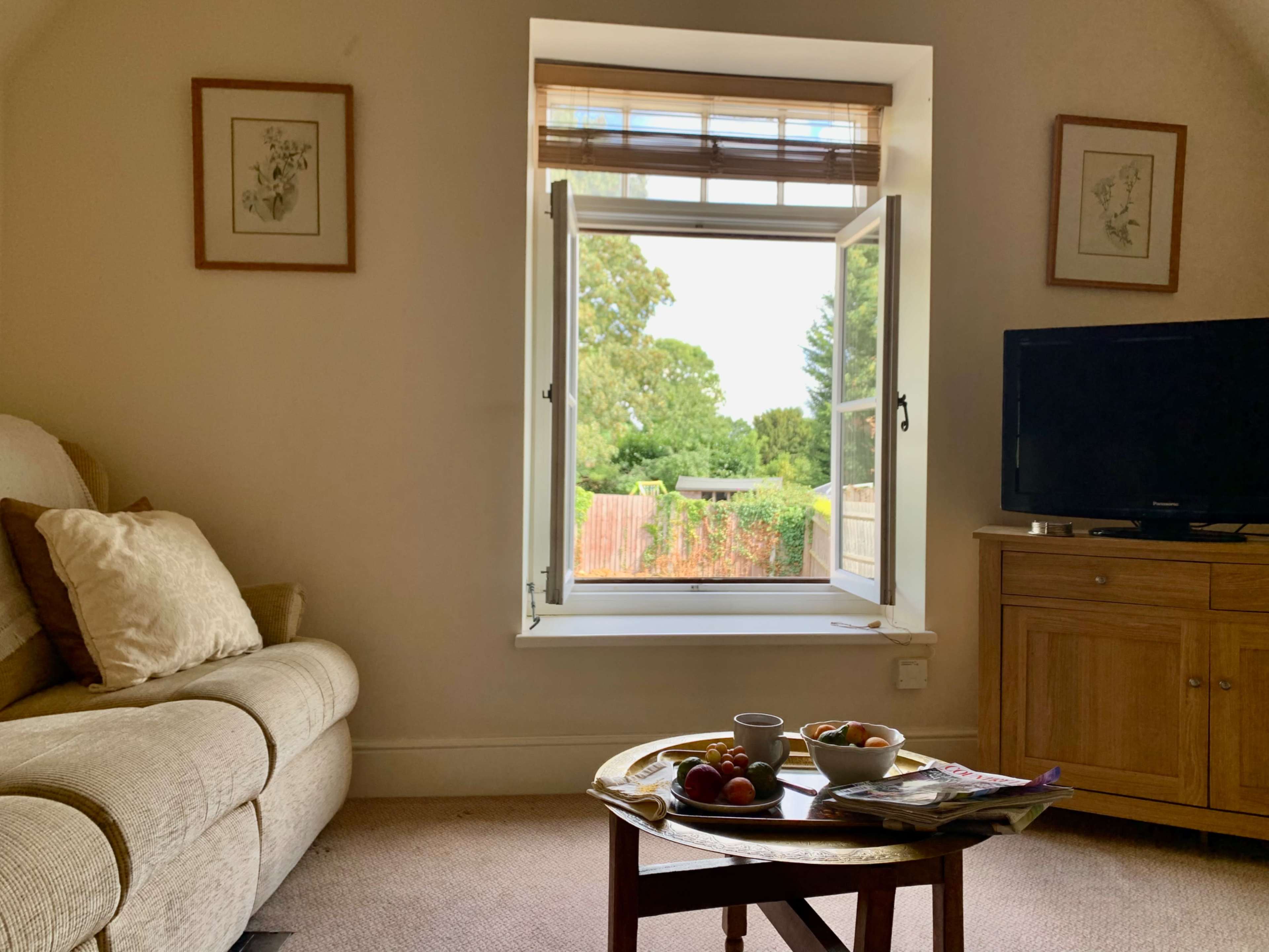 A cozy living room features a sofa, a wooden table with fruits, and an open window overlooking a garden.