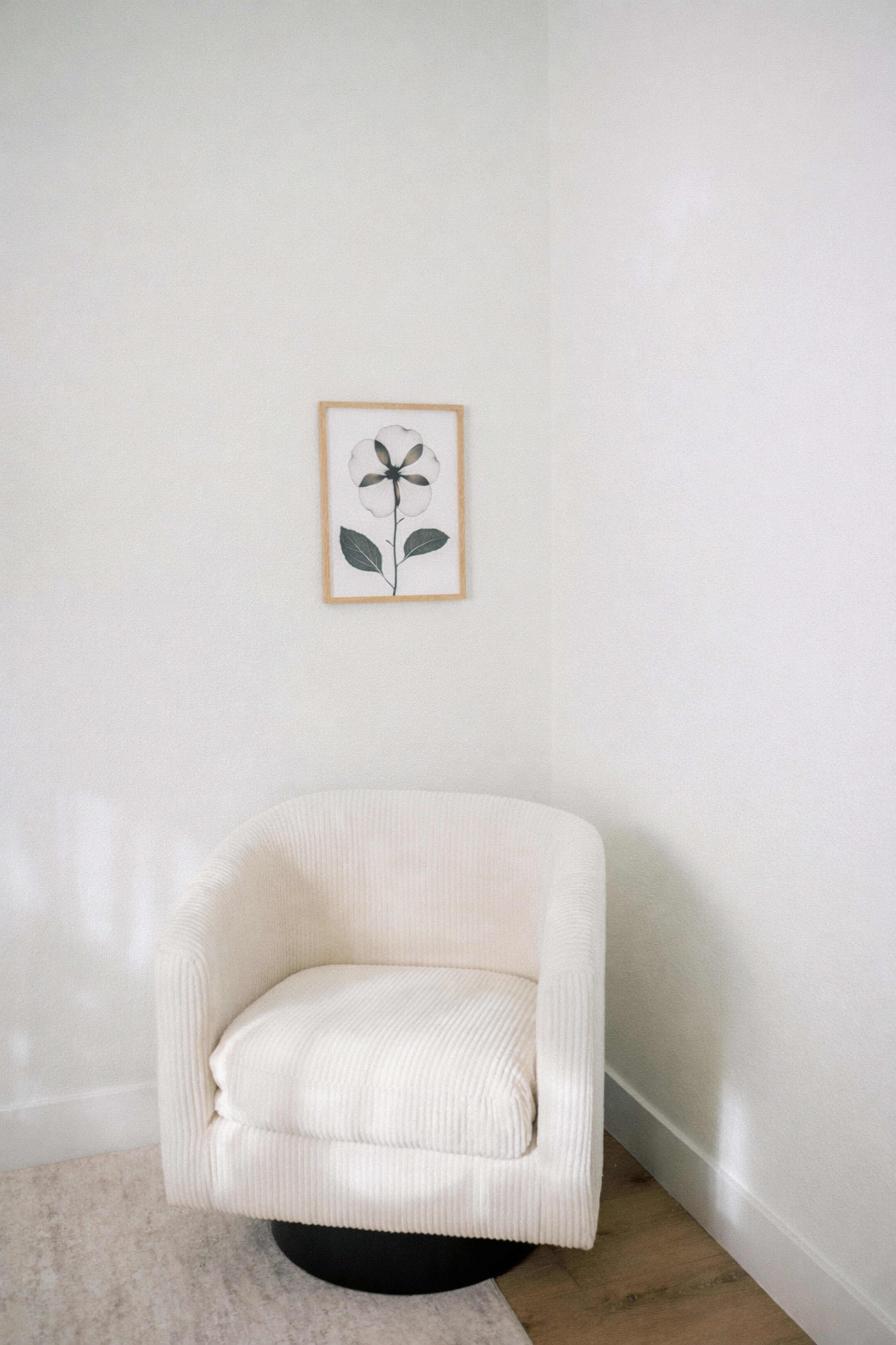 A white upholstered chair is positioned in the corner of a room, next to a framed botanical print on the wall.