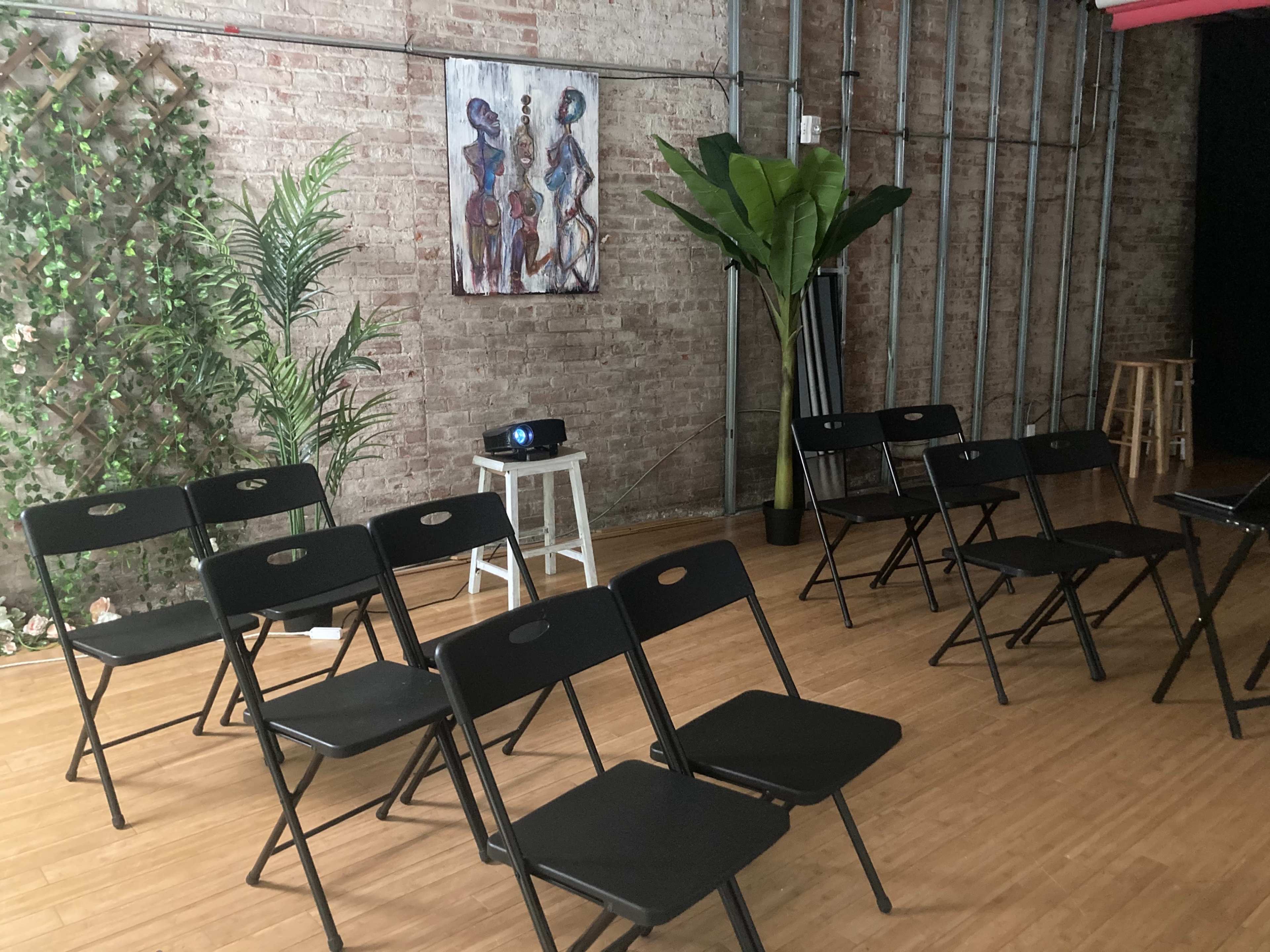 A room with a brick wall, featuring several black folding chairs arranged in rows, a small projector on a white stool, and a painting on the wall alongside a potted plant.
