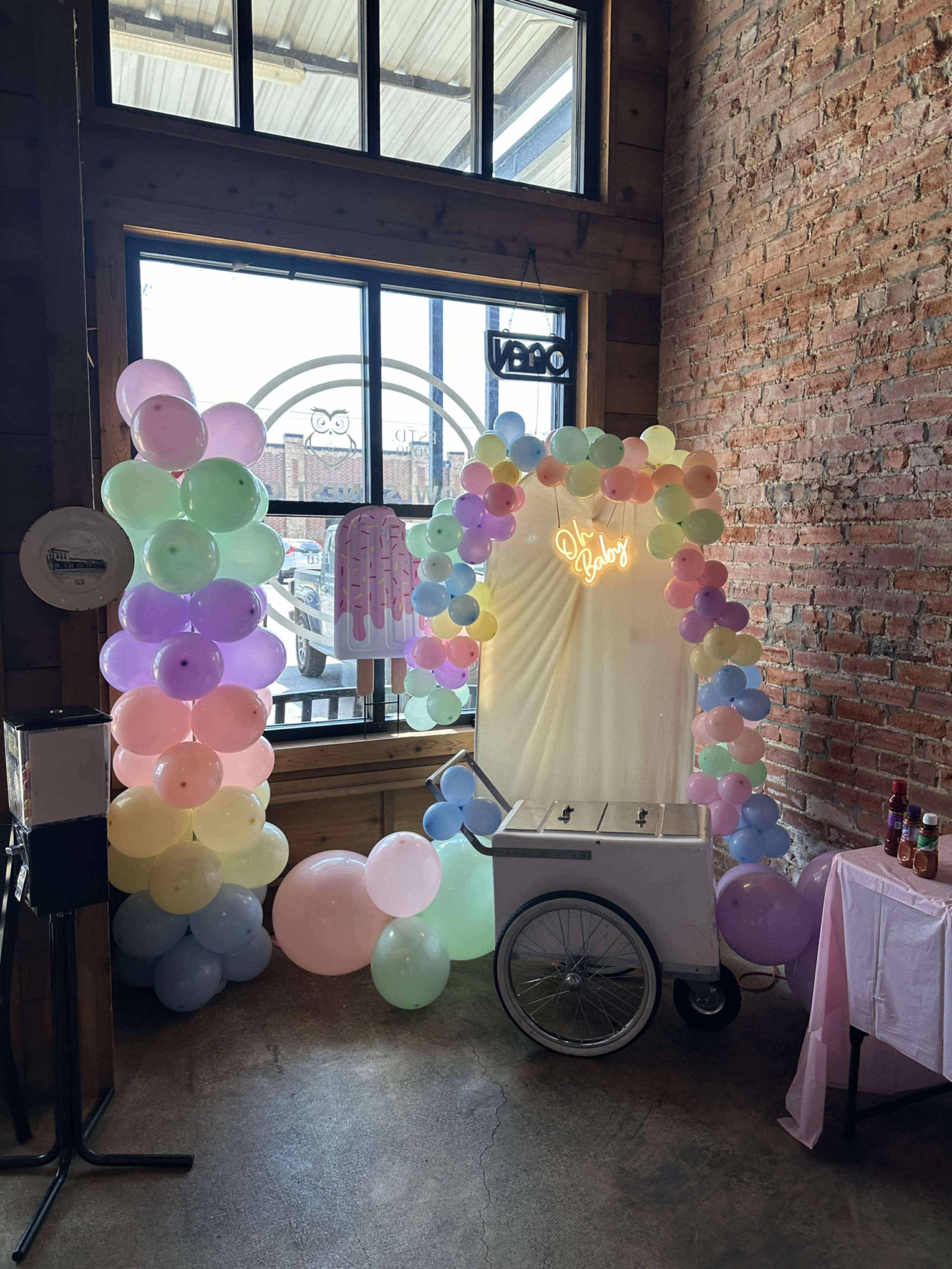 A decorated corner features a vintage ice cream cart surrounded by pastel-colored balloons and a neon sign.