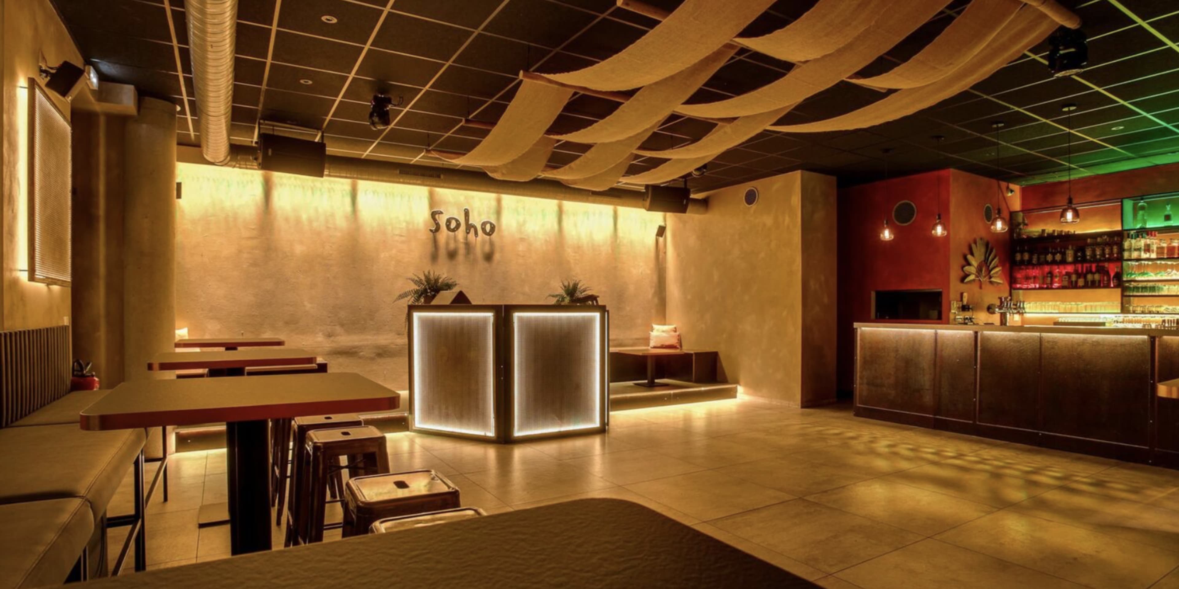 The image shows a modern bar interior with ambient lighting, a rectangular bar counter, and sparse seating arrangements.