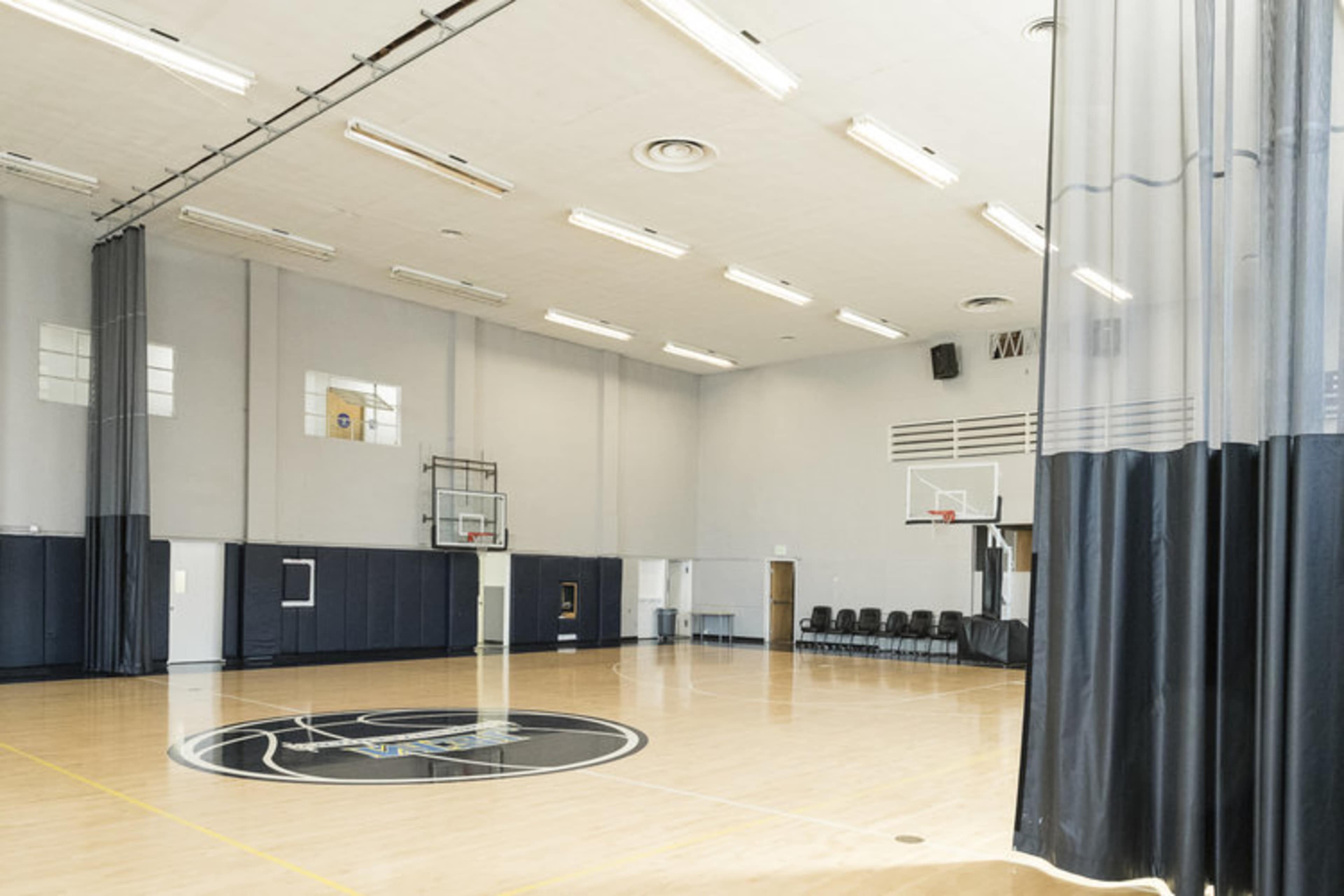 A spacious basketball gymnasium with a polished wooden floor, marked court lines, and a basketball hoop at one end, along with rows of chairs against the wall.