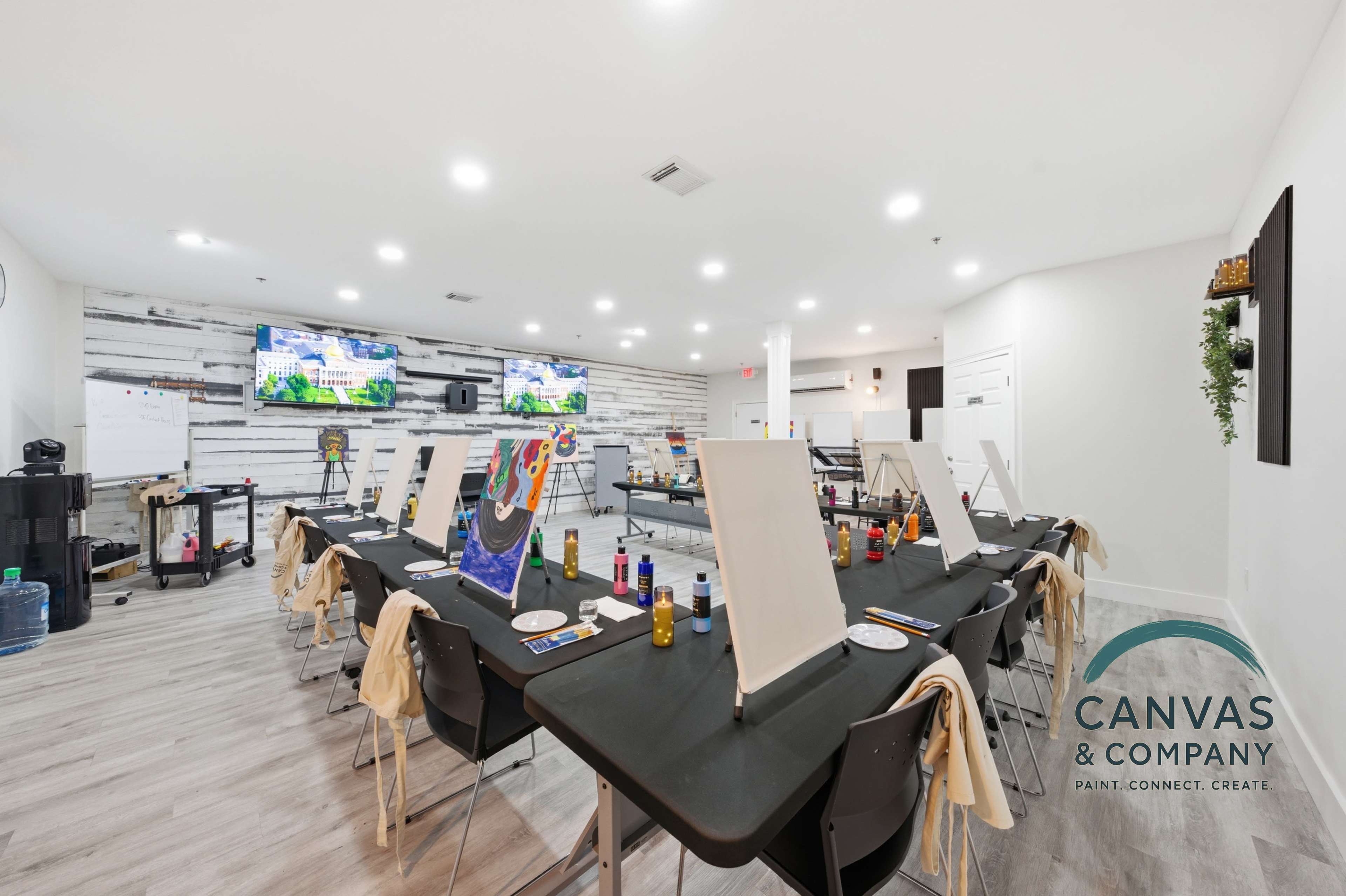The image shows a brightly lit art studio arranged with tables and easels, each set up with art supplies for a painting class.