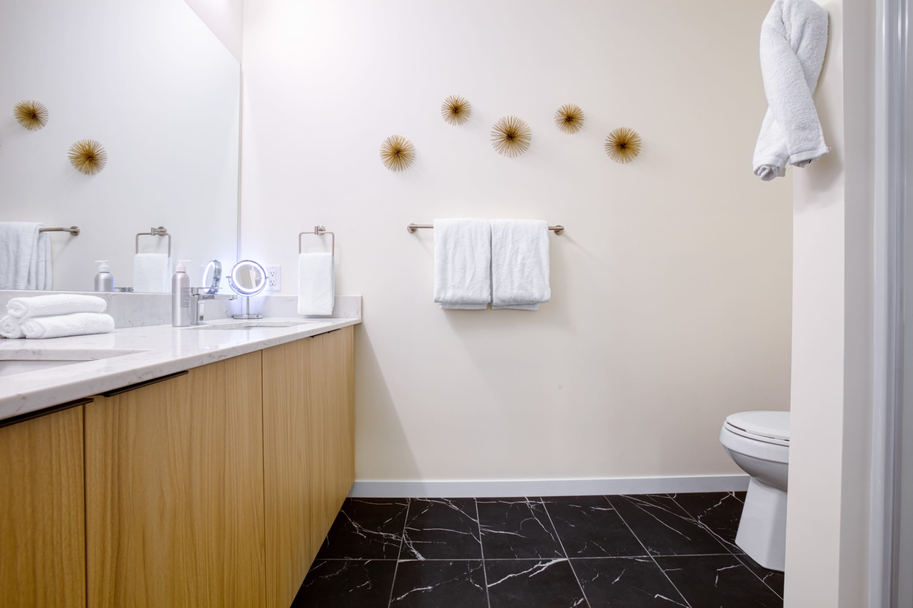 A modern bathroom featuring a white countertop with toiletries, two rolled towels on the counter, a toilet, and decorative wall art above the towel rack.