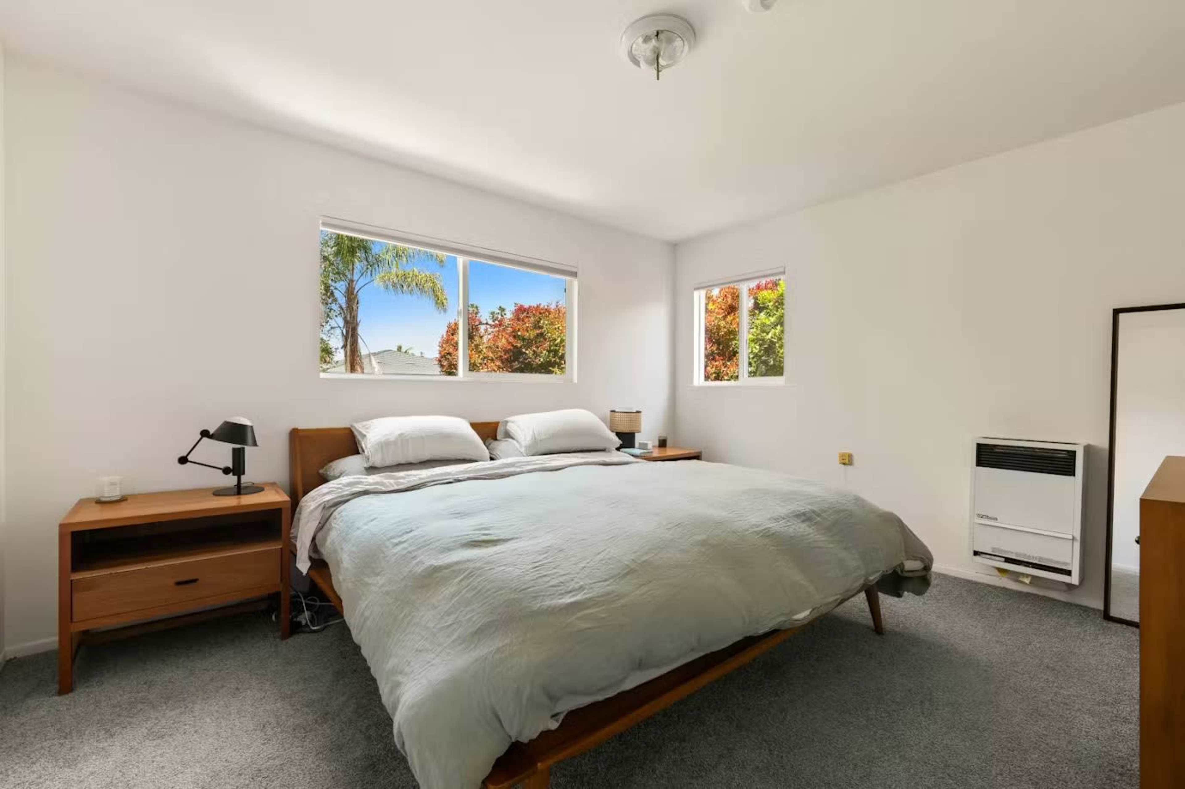 A minimalist bedroom features a large bed with a light gray blanket, two windows allowing natural light, and simple wooden furniture.