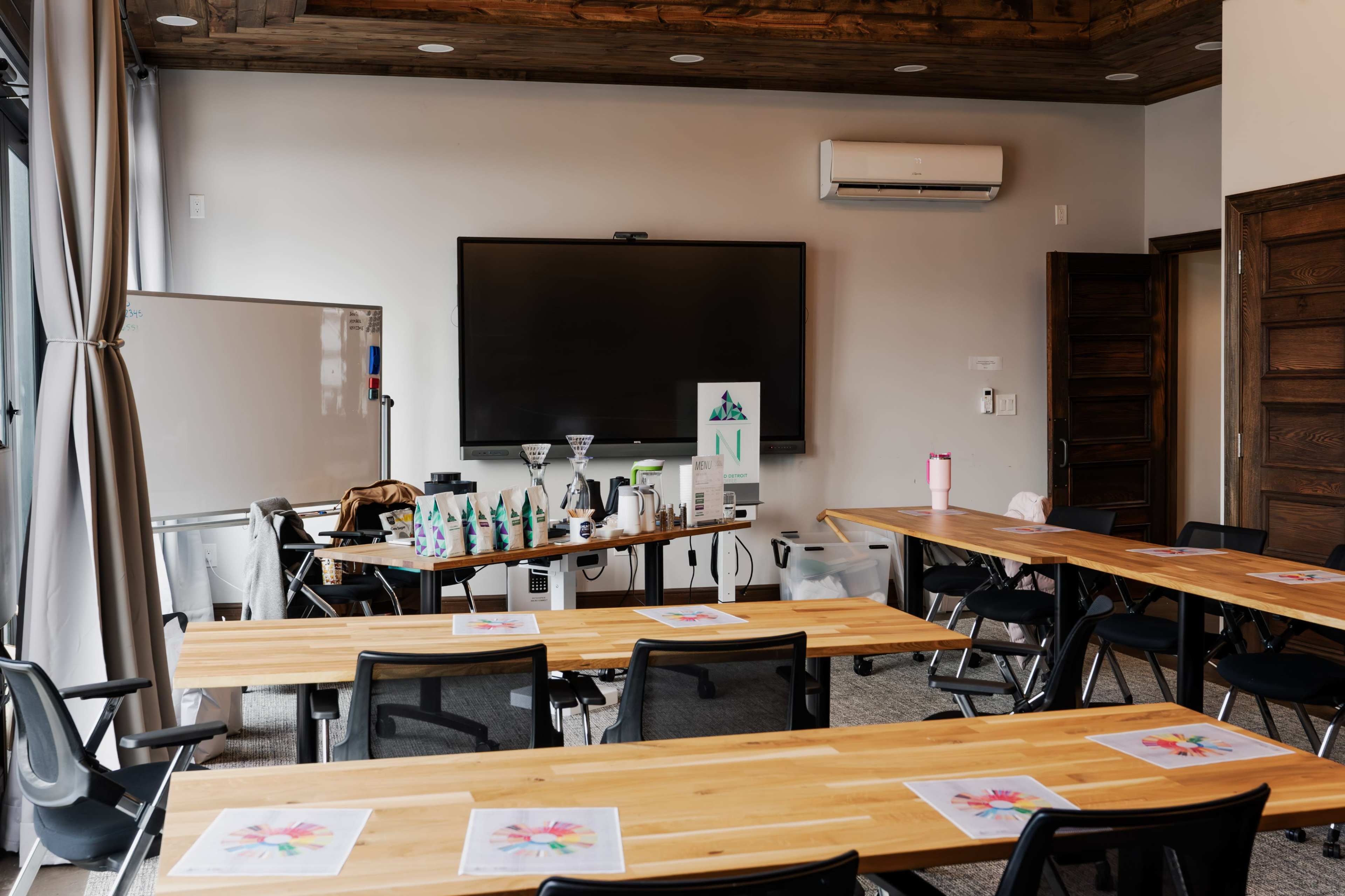 A spacious room with wooden tables and chairs, equipped with a large screen and various presentation materials on a side table.