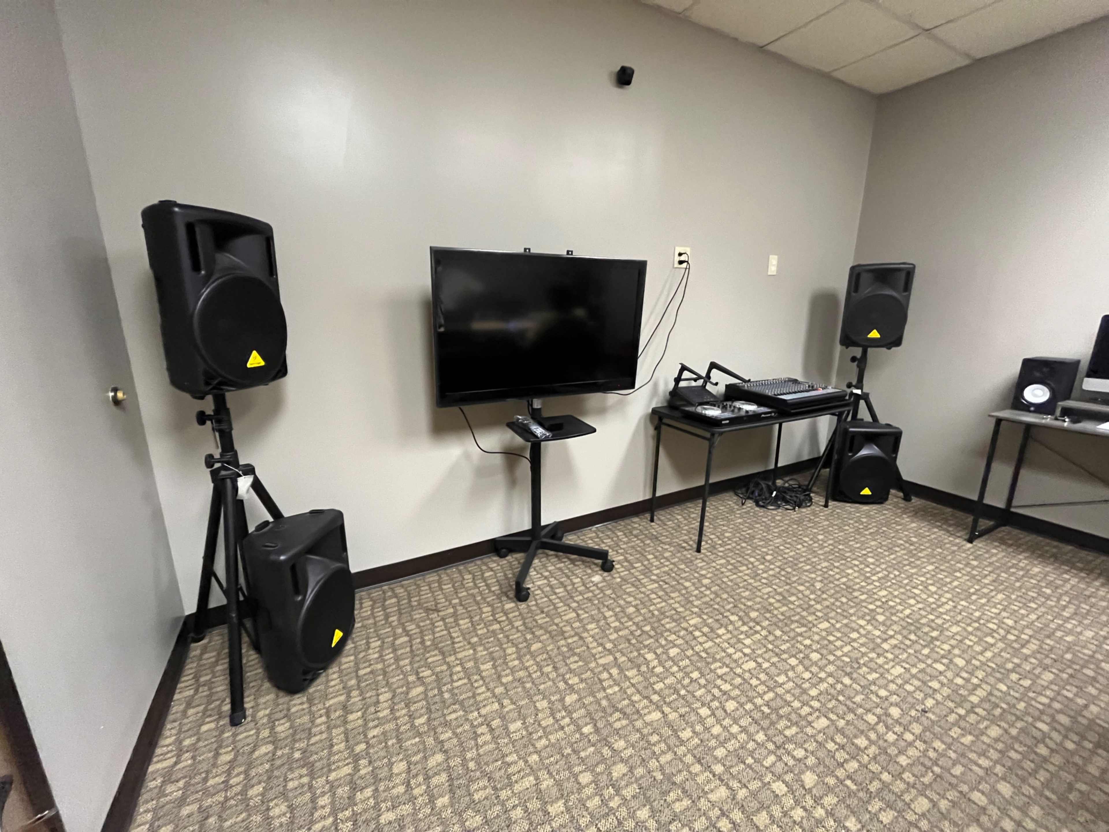 A room with two speakers on stands, a television on a movable stand, and a keyboard or mixer on a table.