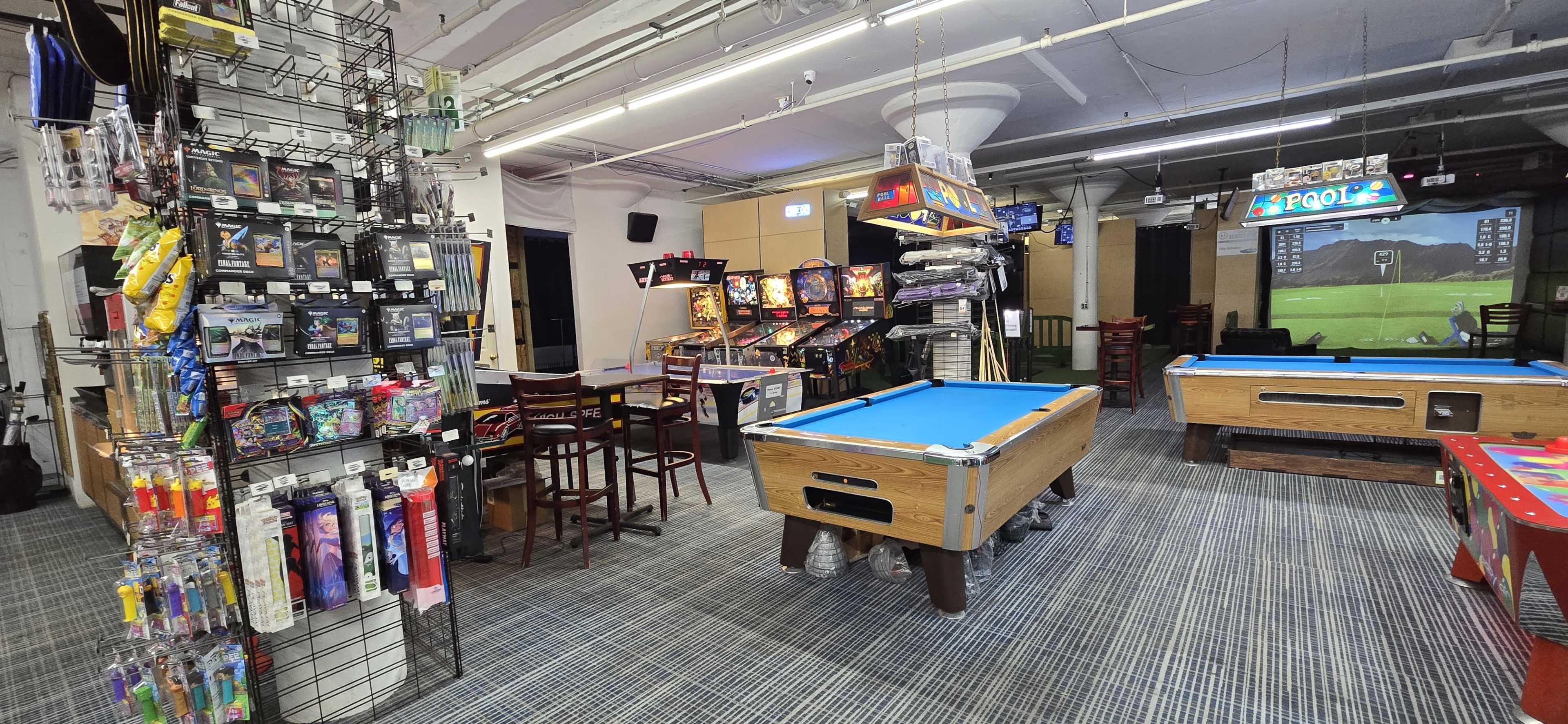 The image shows an entertainment space featuring arcade games, a pool table, and seating areas, with various merchandise displayed on shelves.