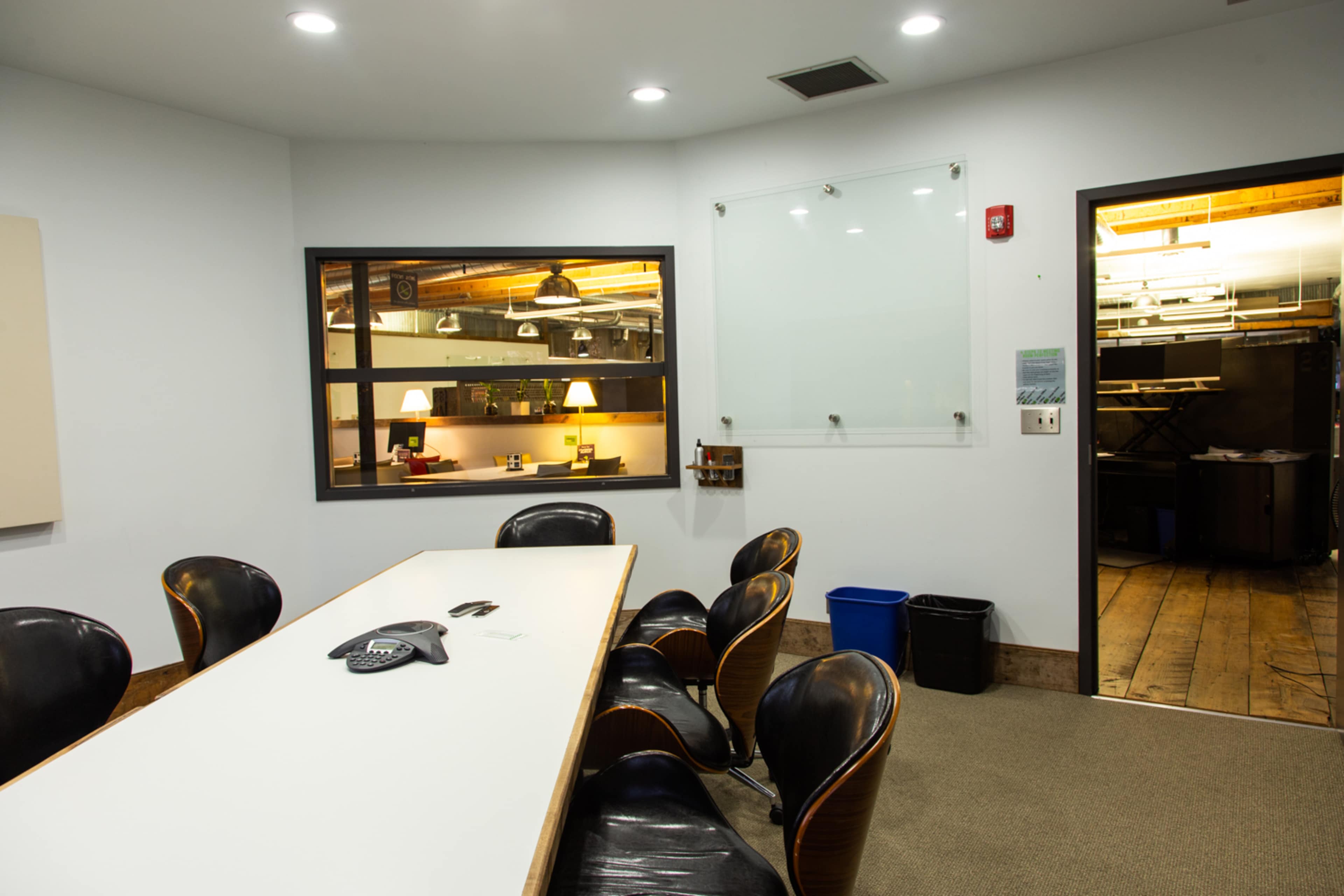 A conference room features a long white table surrounded by black chairs, with a glass marker board on one wall and an entrance to another room.