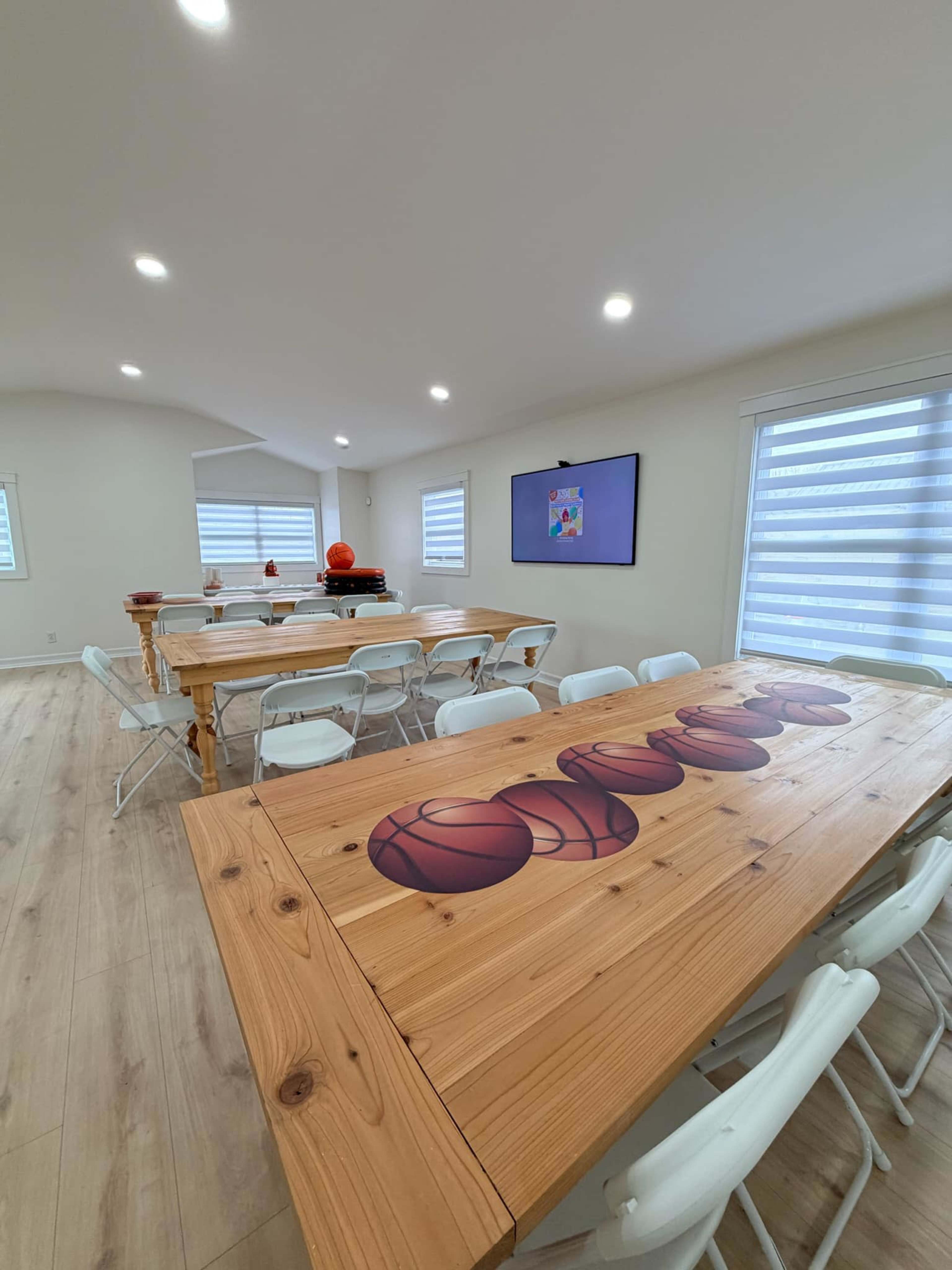 The image shows a well-lit room featuring a large wooden table with basketball designs, surrounded by white folding chairs and a screen mounted on the wall.