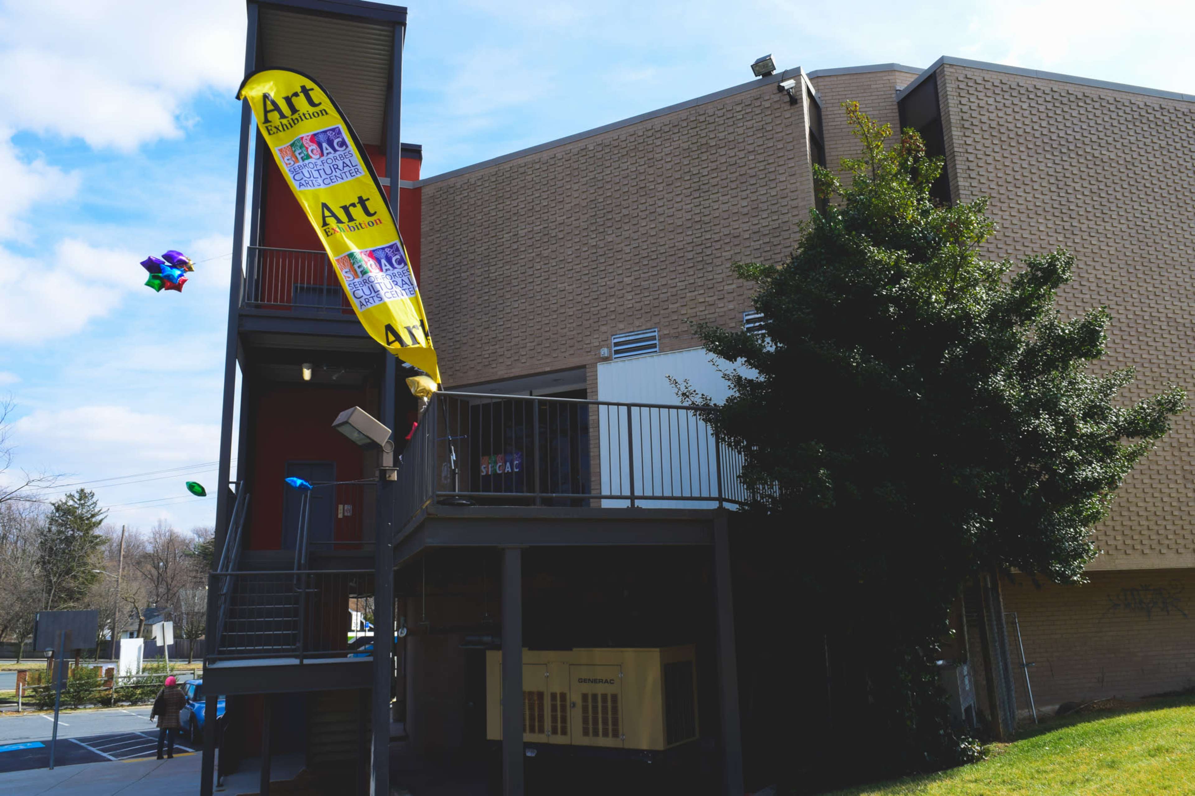 A building with a yellow banner promoting an art exhibition stands next to a small outdoor area with balloons floating nearby.