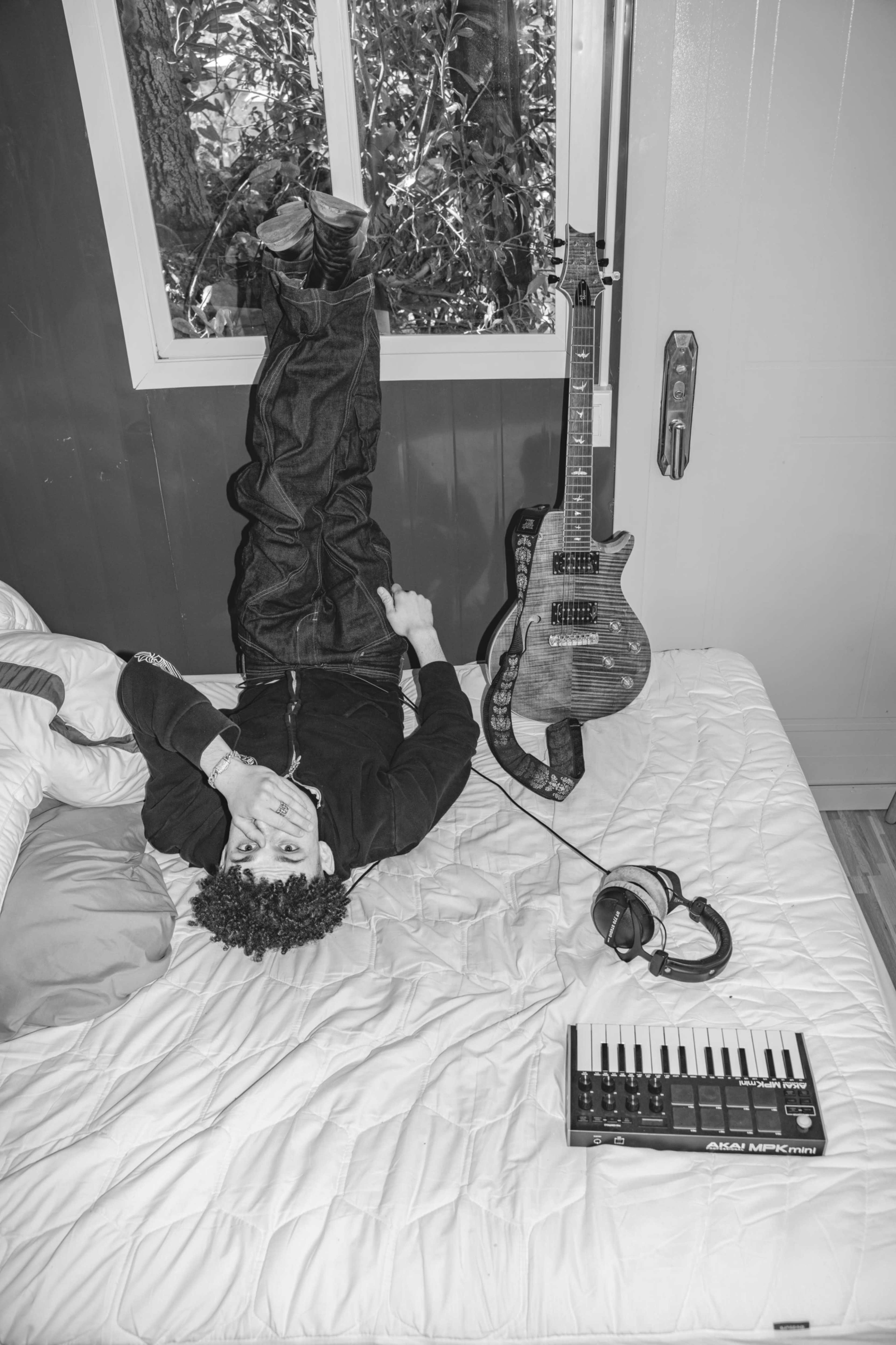 A person lies on a bed with their legs against a wall, next to a guitar and a keyboard, in a room with a window.