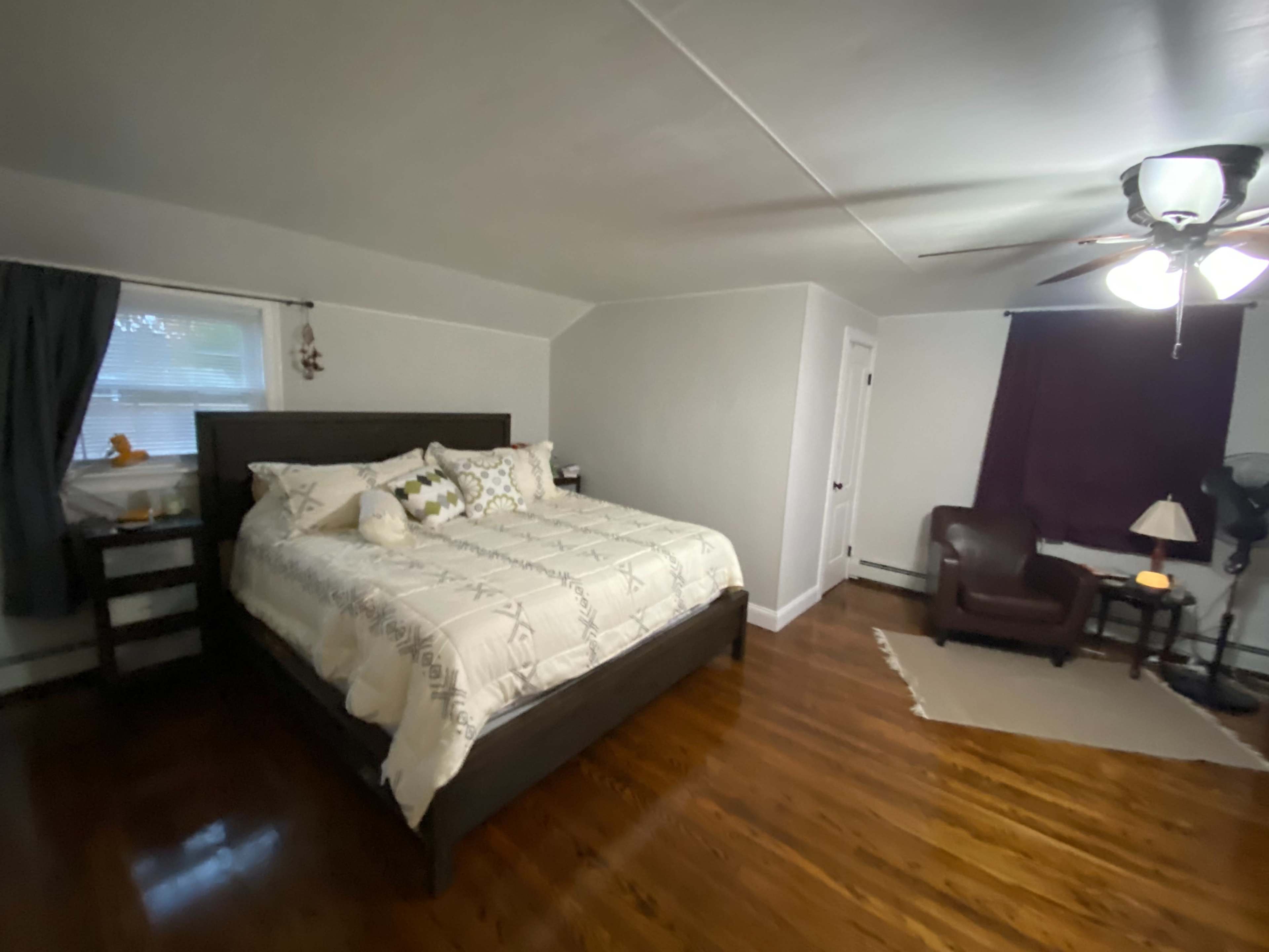 A spacious bedroom features a large bed, a brown armchair, and a window with sheer curtains, along with wood flooring and a ceiling fan.