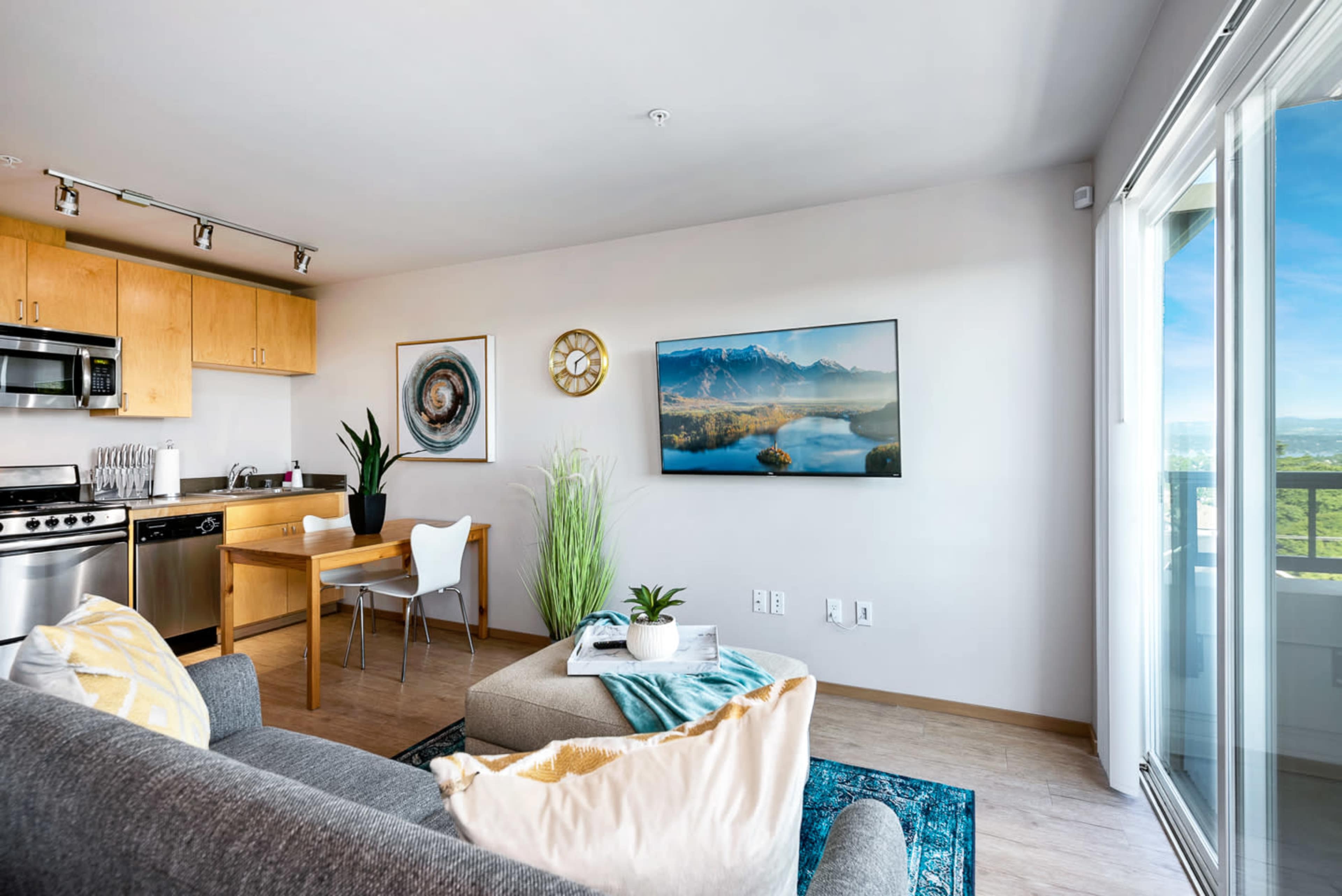 The image shows a modern living space with a kitchen area, a chair and dining table, a couch, and a large window displaying a scenic view.