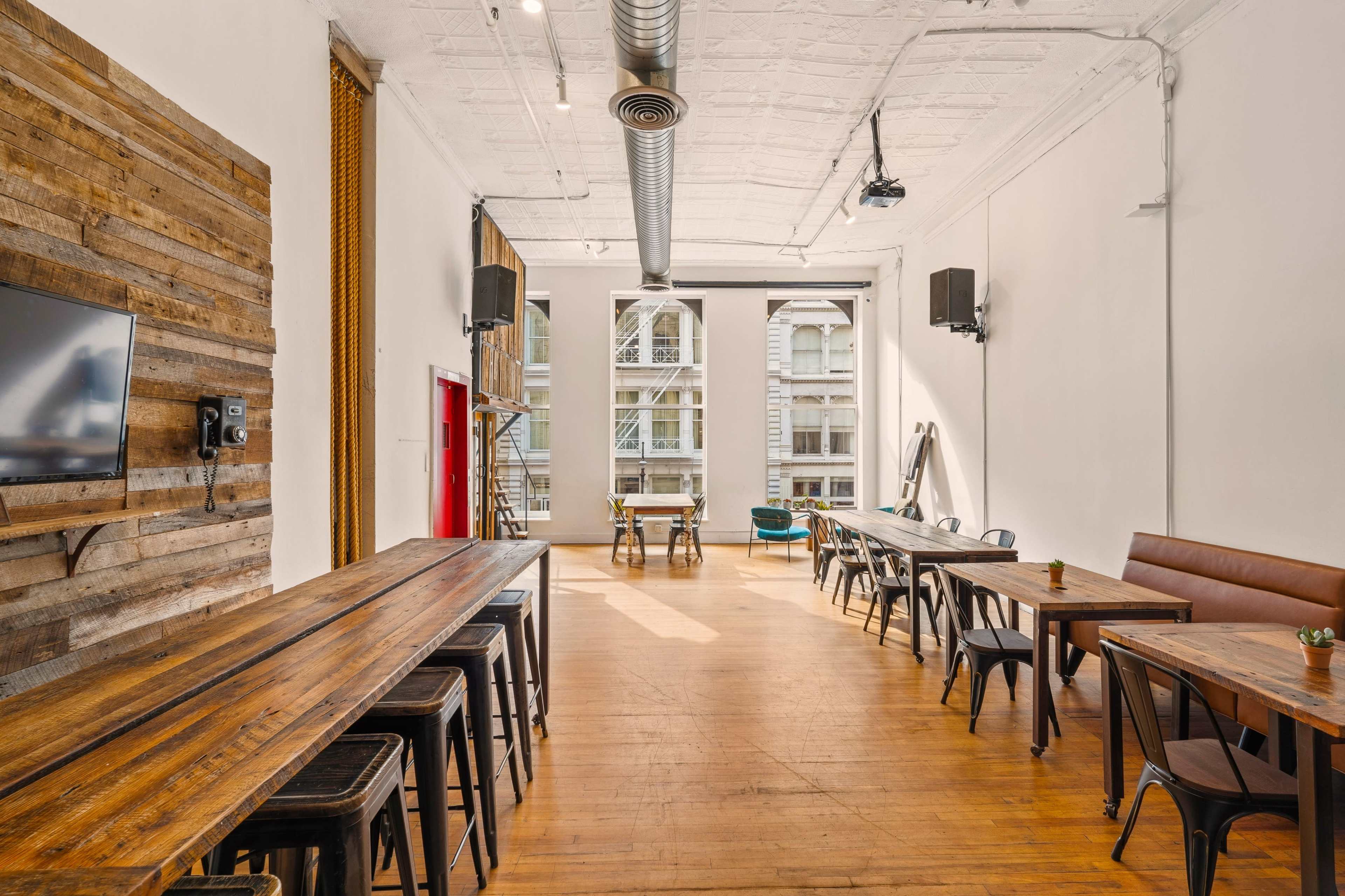 The room features several long wooden tables and chairs arranged in an open layout with large windows providing natural light.