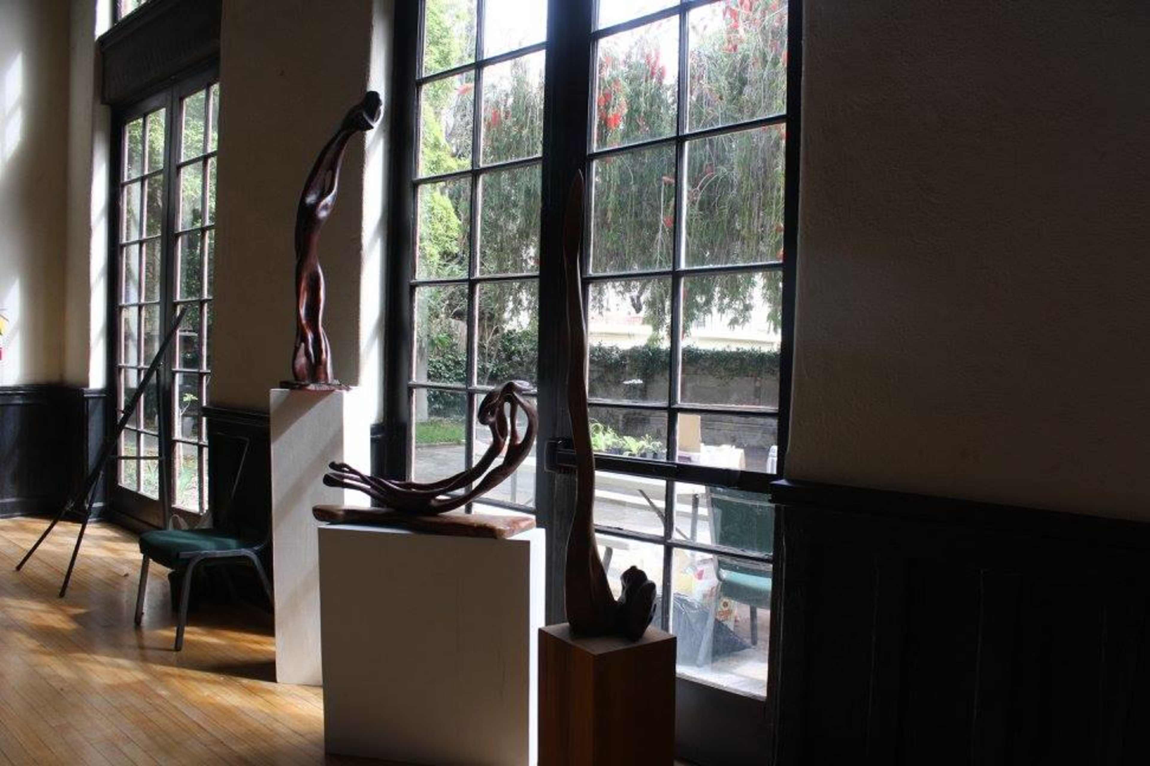 An interior space featuring three wooden sculptures displayed on pedestals beside large windows with outdoor greenery visible.