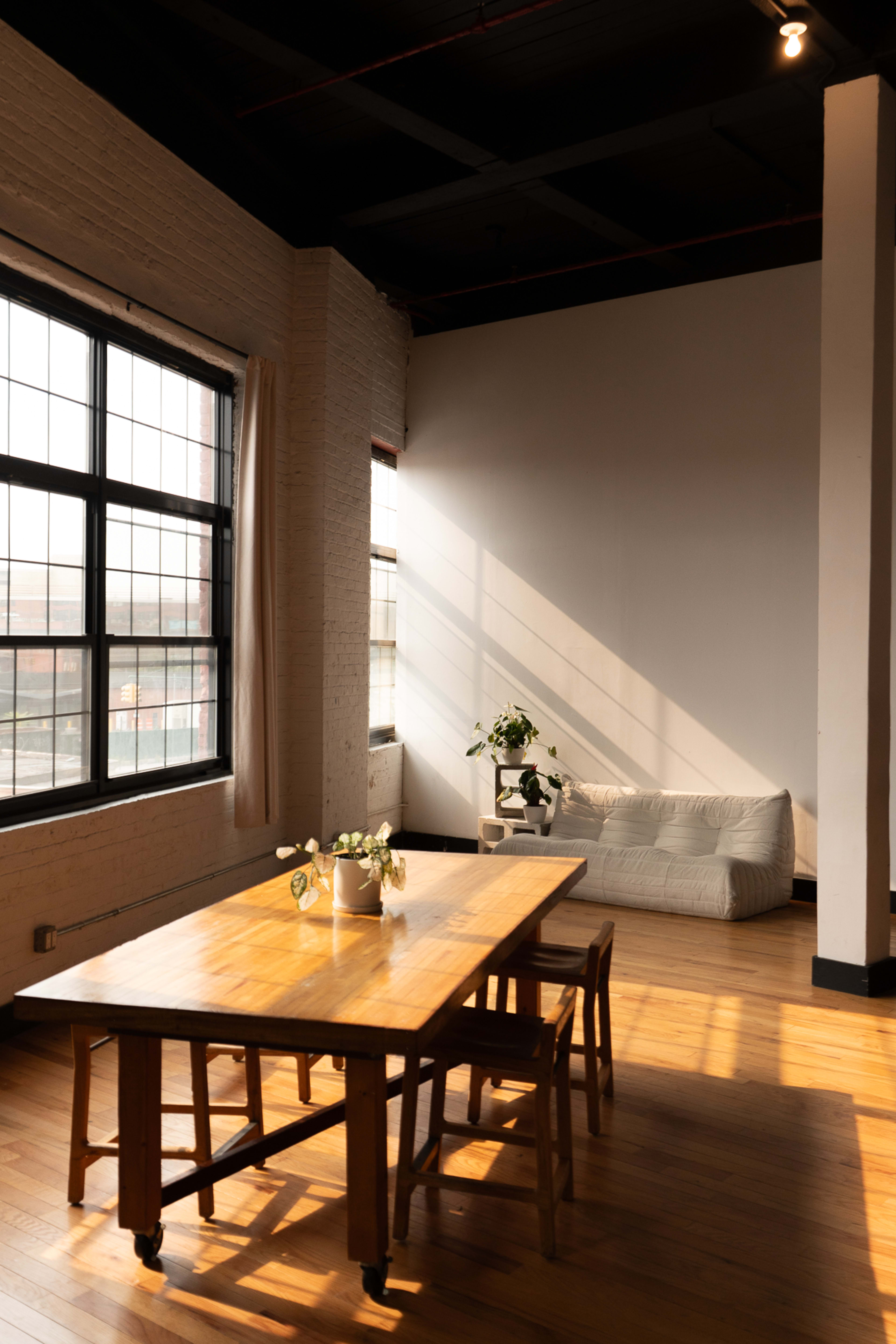 The image shows a minimalist room with large windows, featuring a wooden table and chairs, a couch, and a small plant on the table.