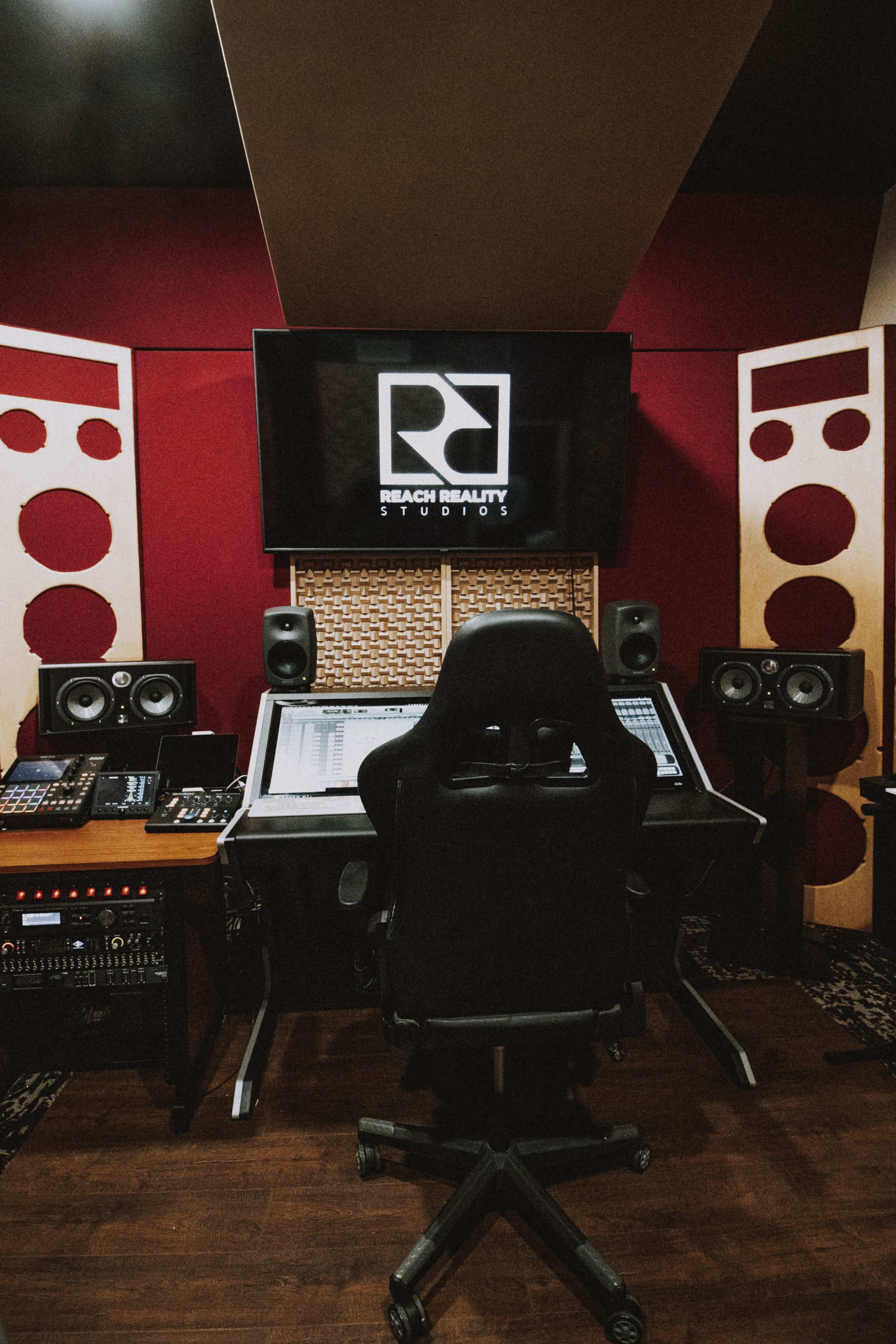 A music studio with a large mixing console, speakers, and a central black chair facing a screen displaying the studio logo.