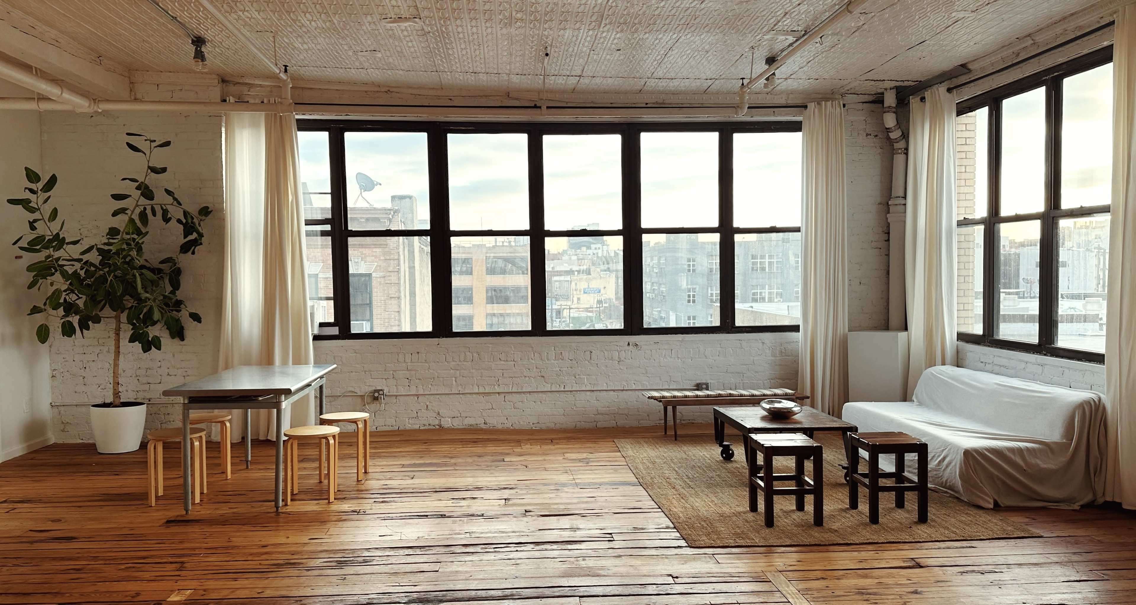 A spacious, sunlit room with large windows, wooden floors, a simple sofa covered in white fabric, a dining table, and various small furniture pieces.