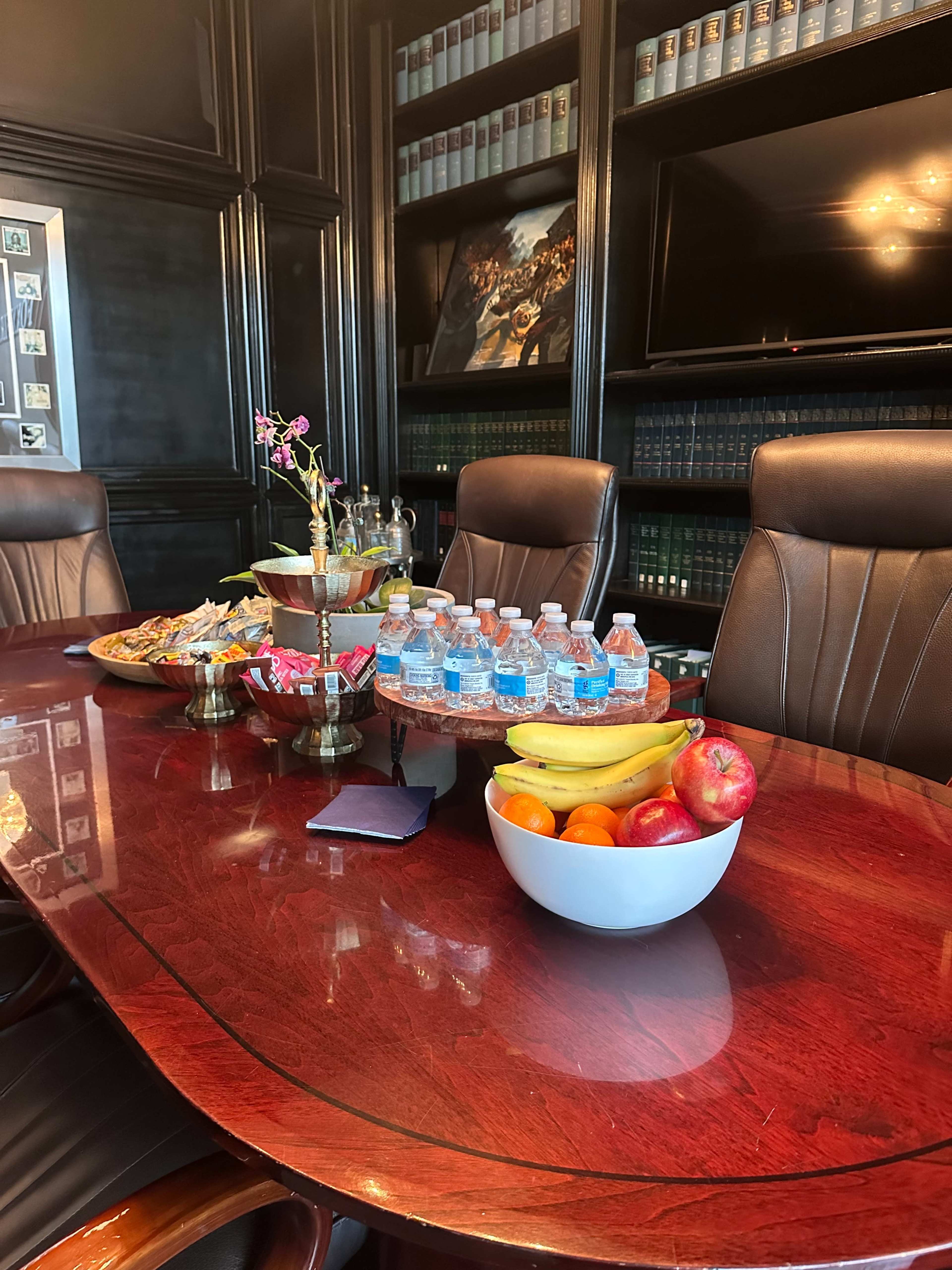 The image shows a conference room table set with bottled water, a bowl of assorted fruits, snacks, and a decorative centerpiece, surrounded by leather chairs and shelves of books in the background.