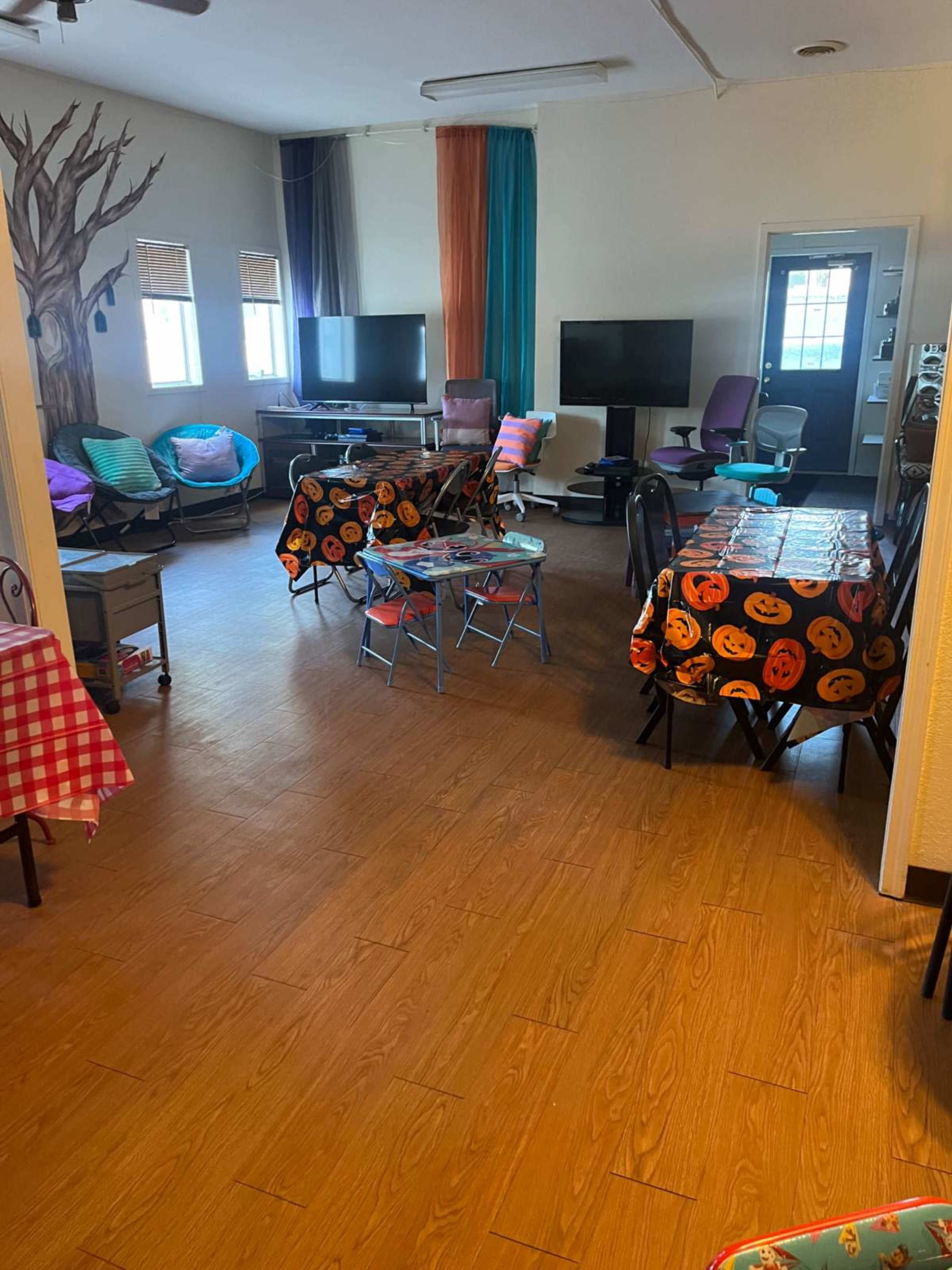 The image shows a spacious room with several tables covered in patterned tablecloths, multiple chairs, and two TVs positioned against the walls.