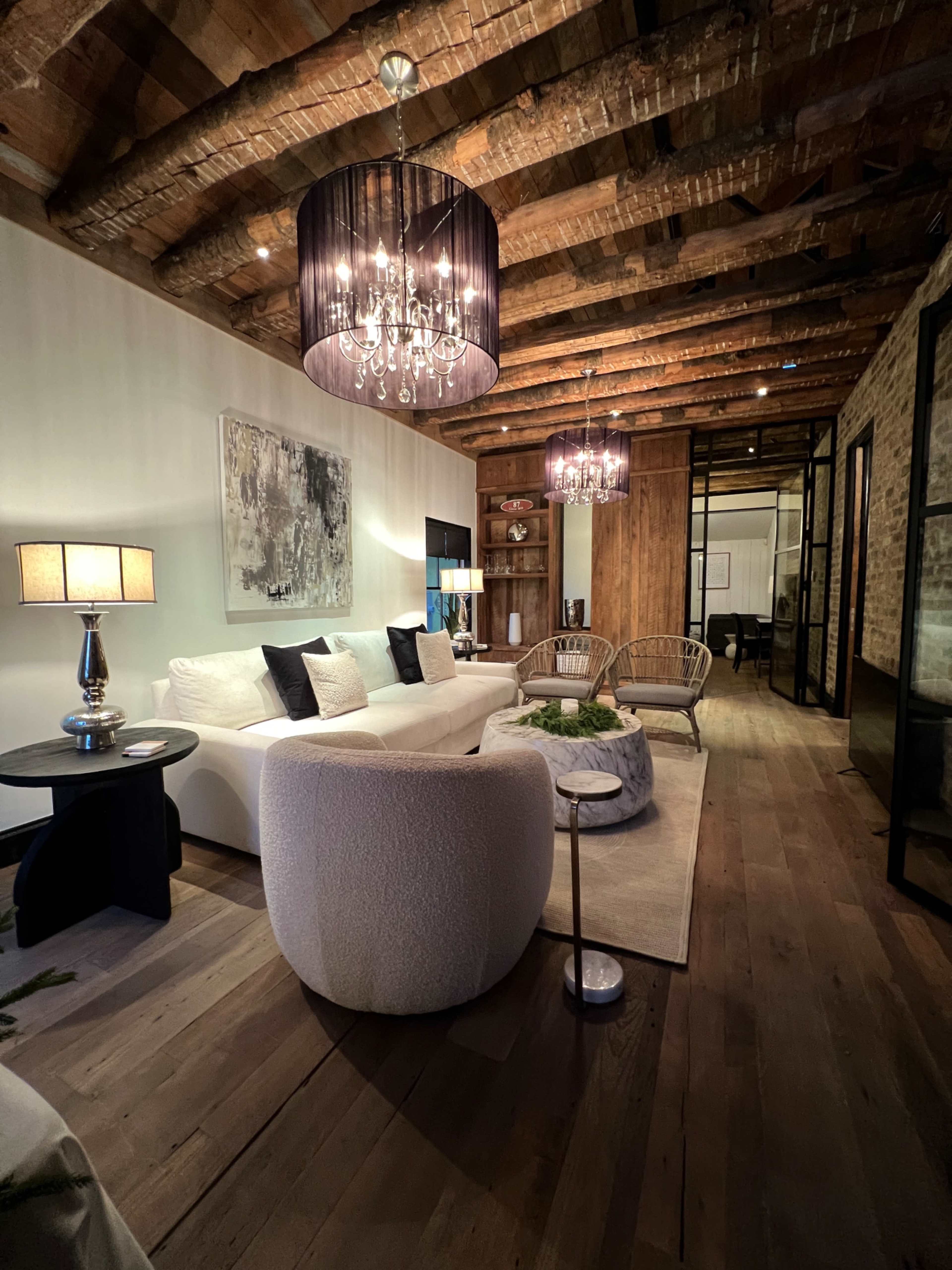 The image shows a modern living room with a white sofa, two rattan chairs, a coffee table, and exposed wooden beams on the ceiling.