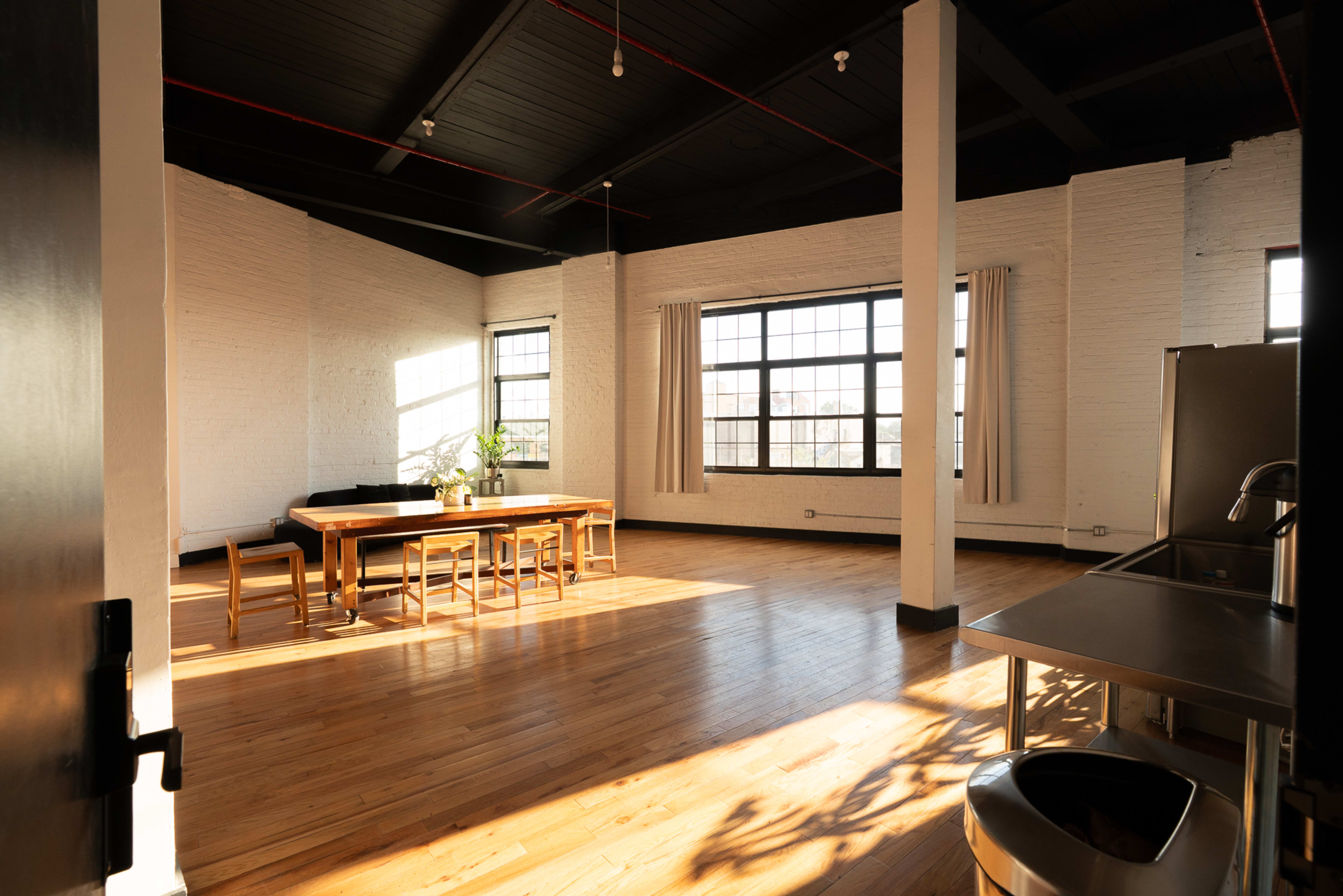 The image shows a spacious, modern room with large windows, a wooden dining table surrounded by chairs, and a kitchenette in the corner.