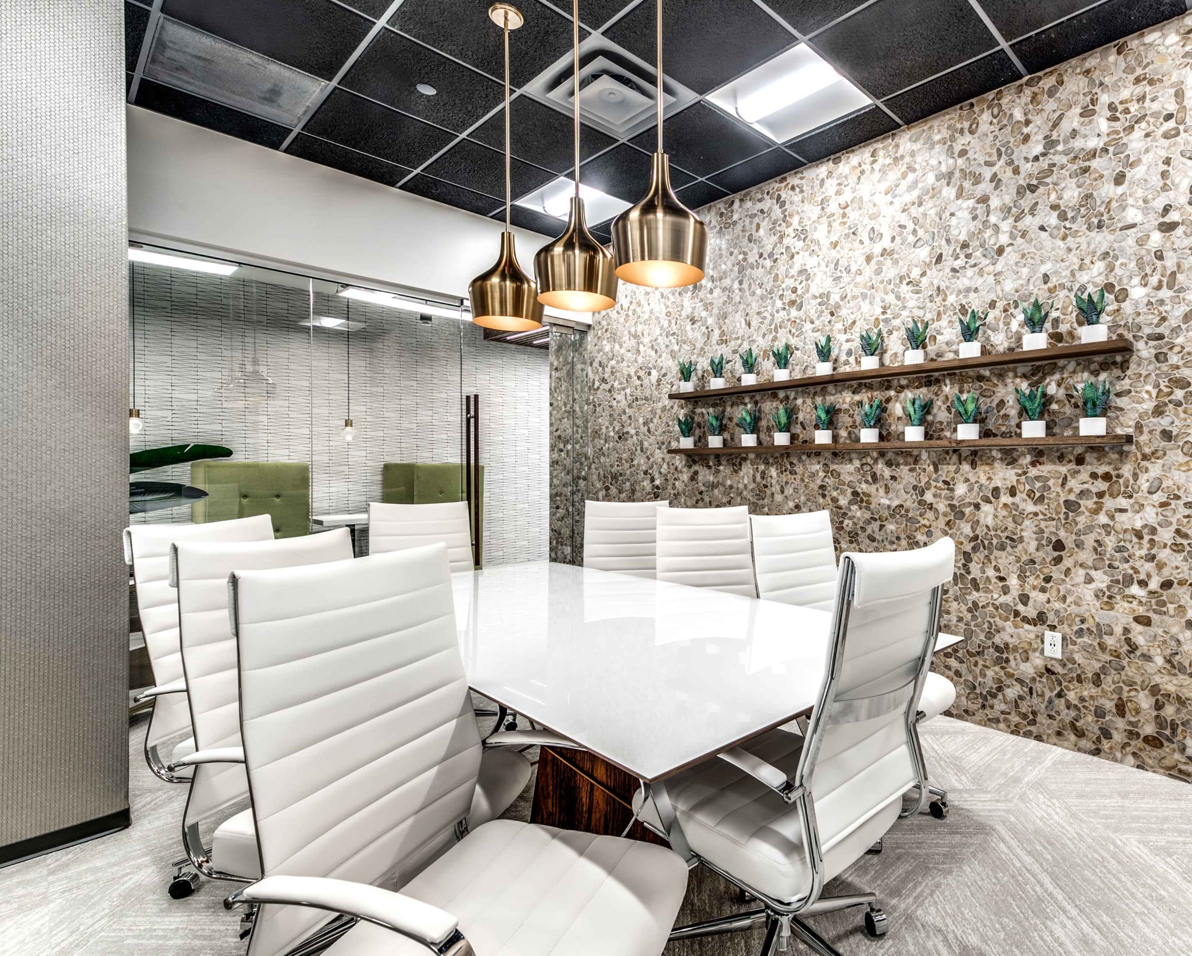 A modern conference room features a white marble table surrounded by sleek white chairs, with a stone accent wall decorated with small potted plants and pendant lighting overhead.