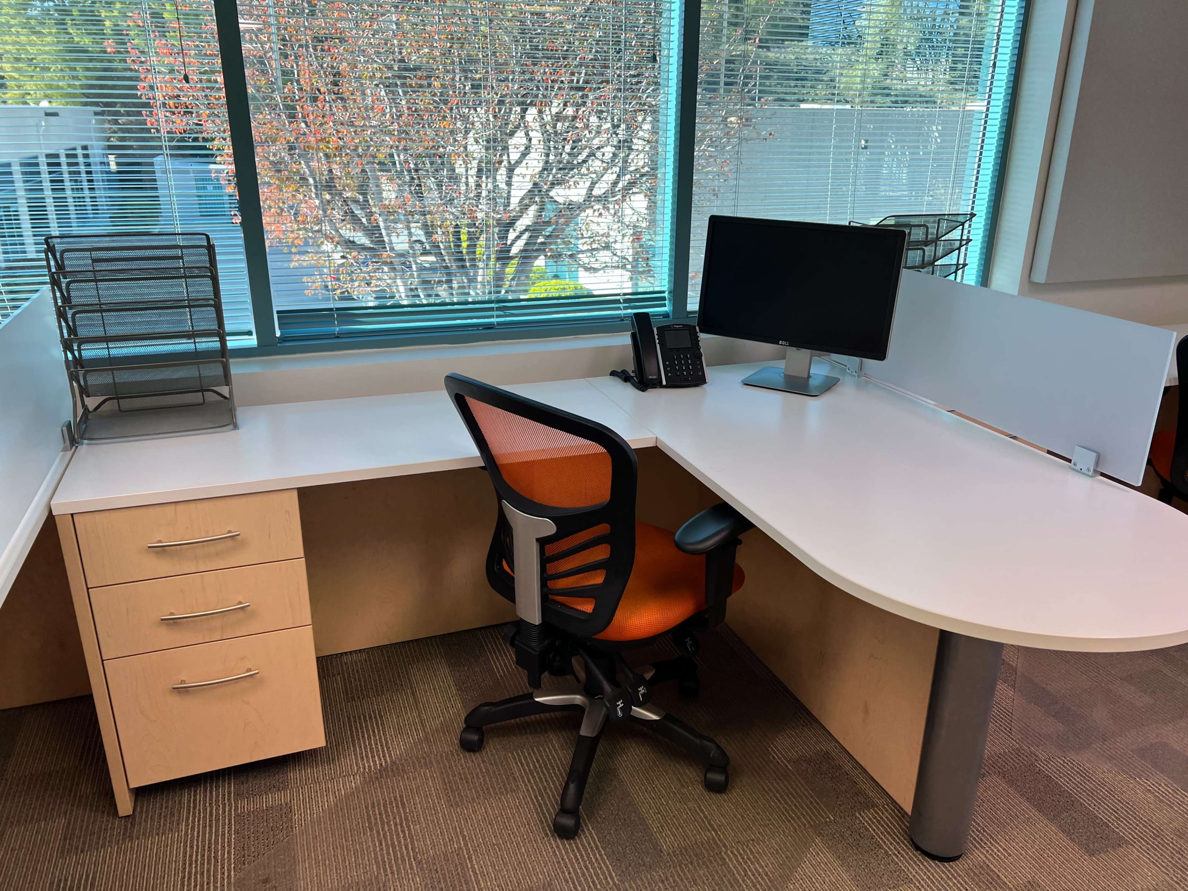 A modern office workspace with a white desk, an orange office chair, a computer monitor, a telephone, and a filing rack, all positioned by a large window.