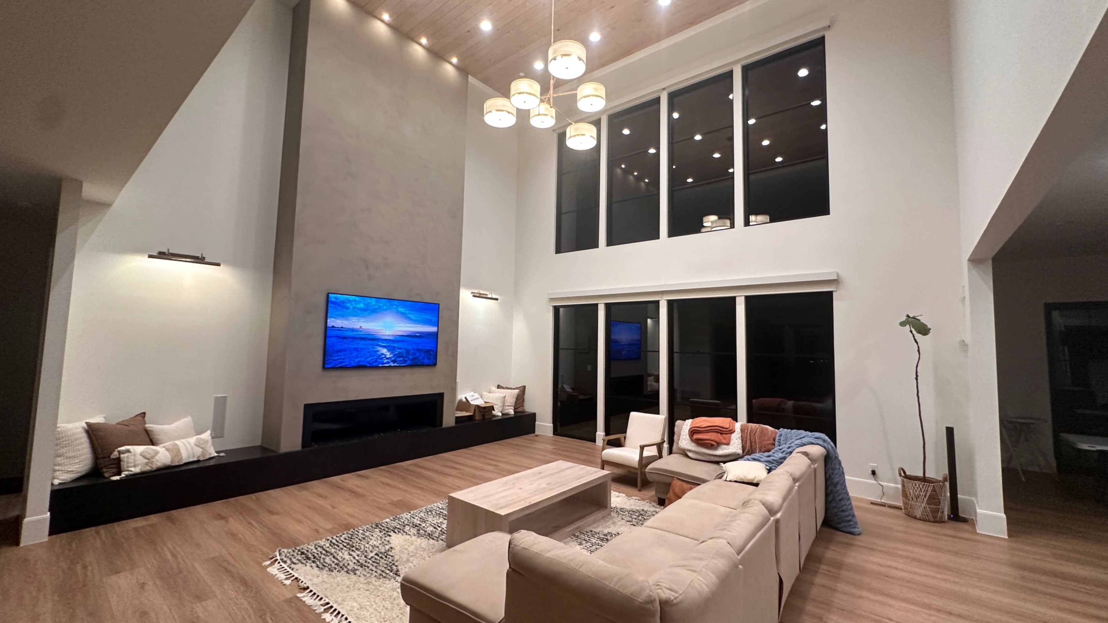 A modern living room features a large sectional sofa, a coffee table, a wall-mounted television displaying a scenic view, large windows, and a high ceiling adorned with multiple light fixtures.