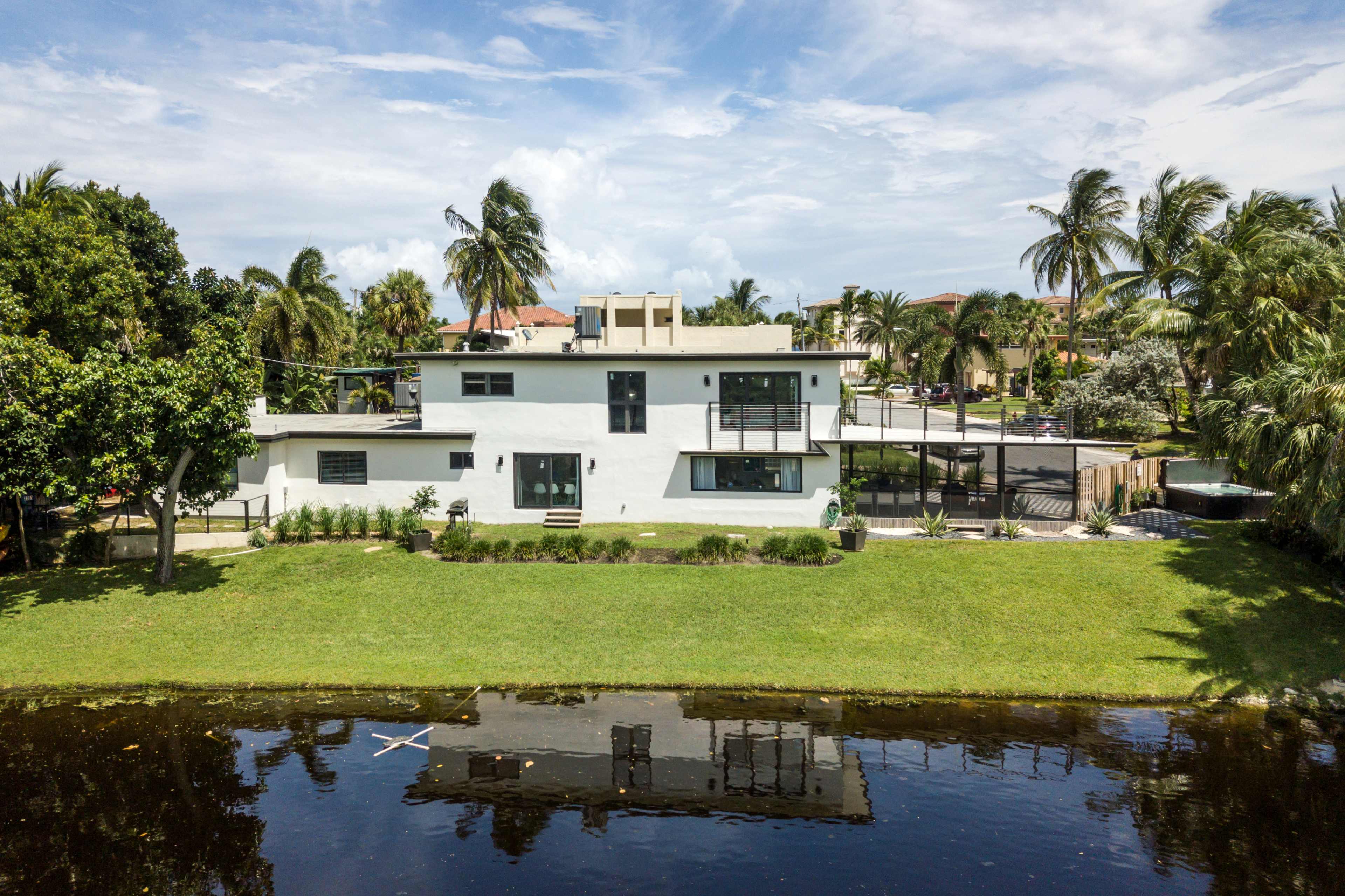 A modern house with large windows and a landscaped yard reflects in a nearby body of water, surrounded by palm trees and residential buildings.