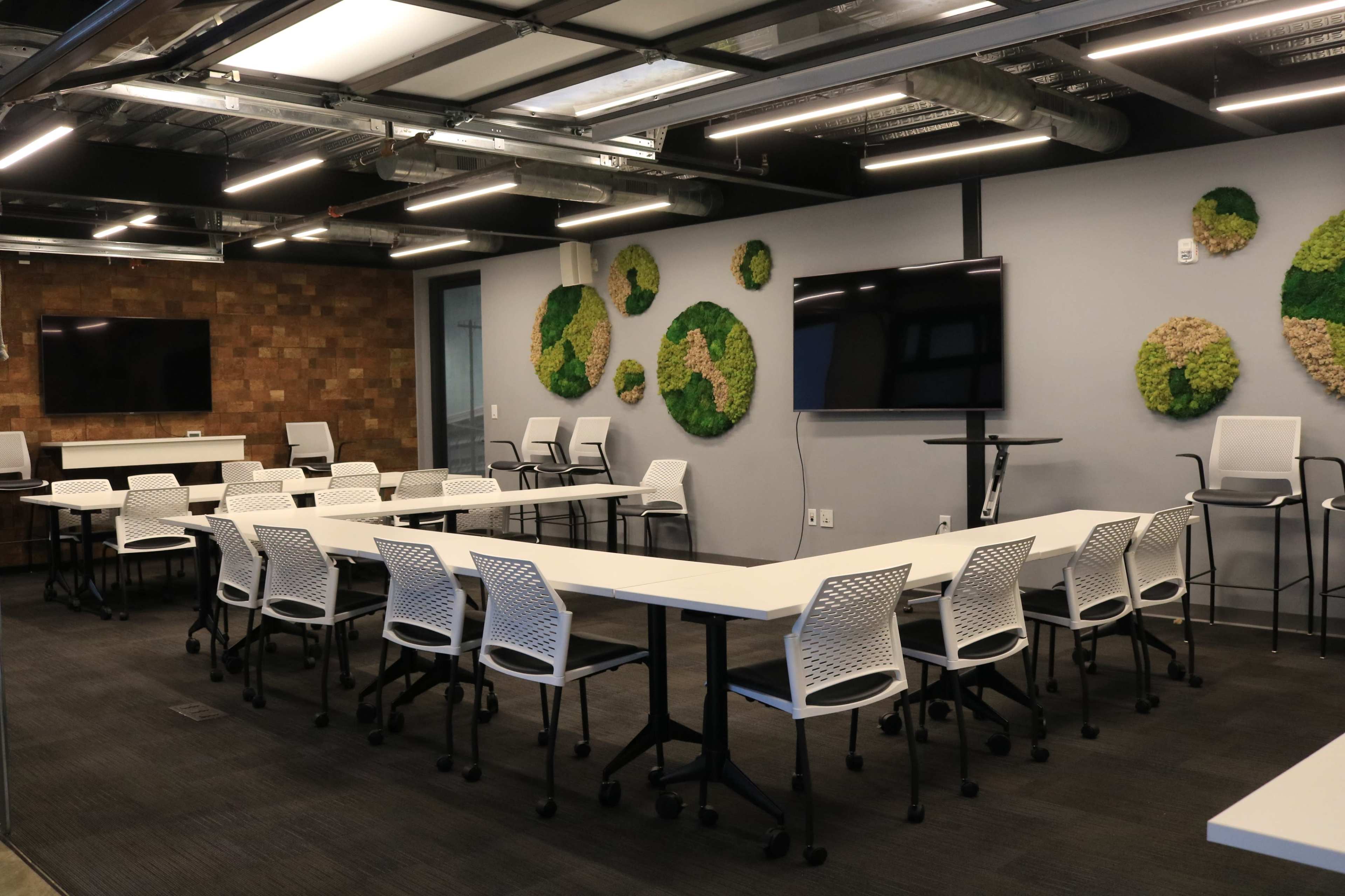 A modern conference room features multiple white tables and chairs arranged for a meeting, with green wall decor and large screens on the walls.
