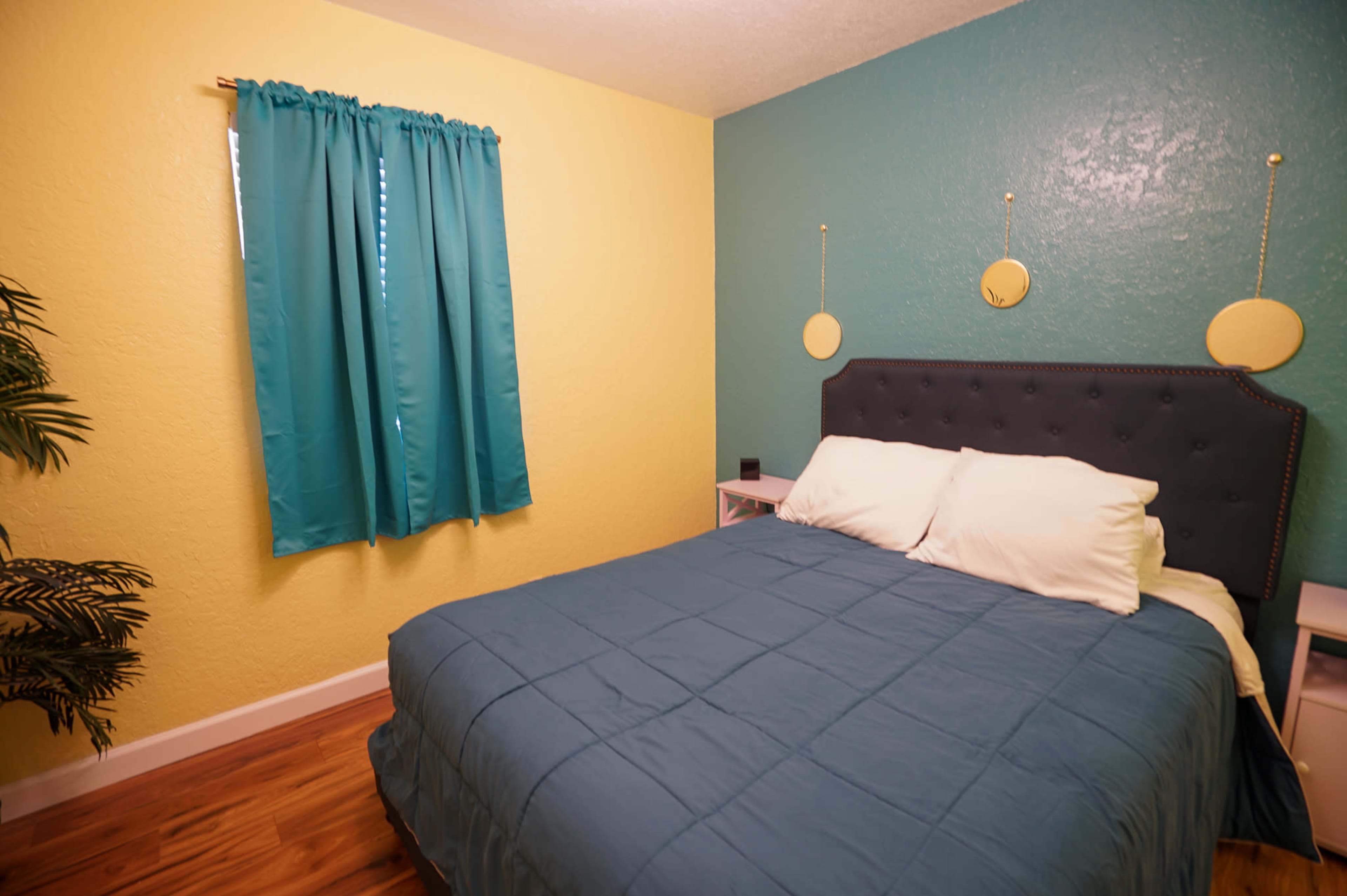 The image shows a small bedroom with a teal bedspread, a bed with white pillows, patterned walls in yellow and teal, and a window with teal curtains.