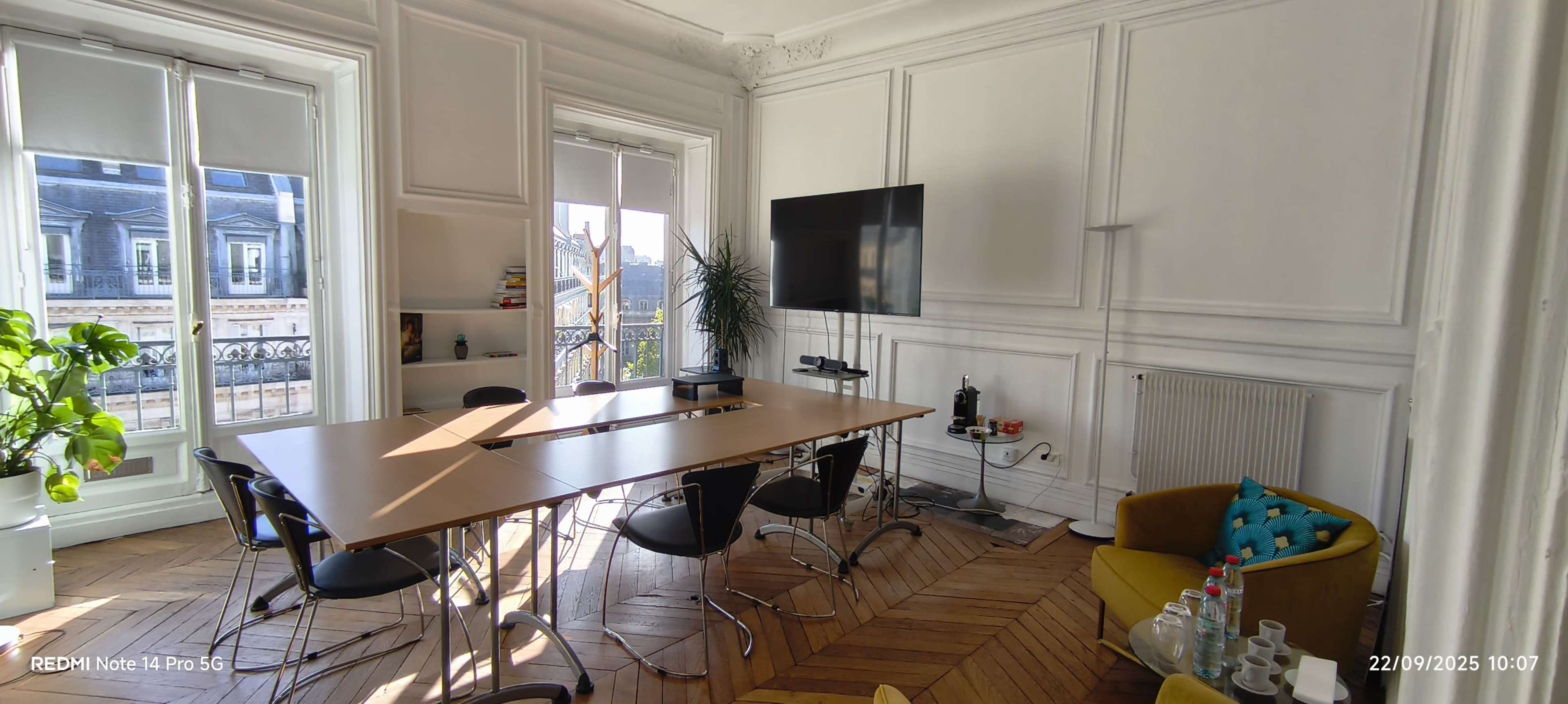 The image shows a well-lit conference room with a long wooden table, several chairs, a large television on the wall, and a plant in the corner.