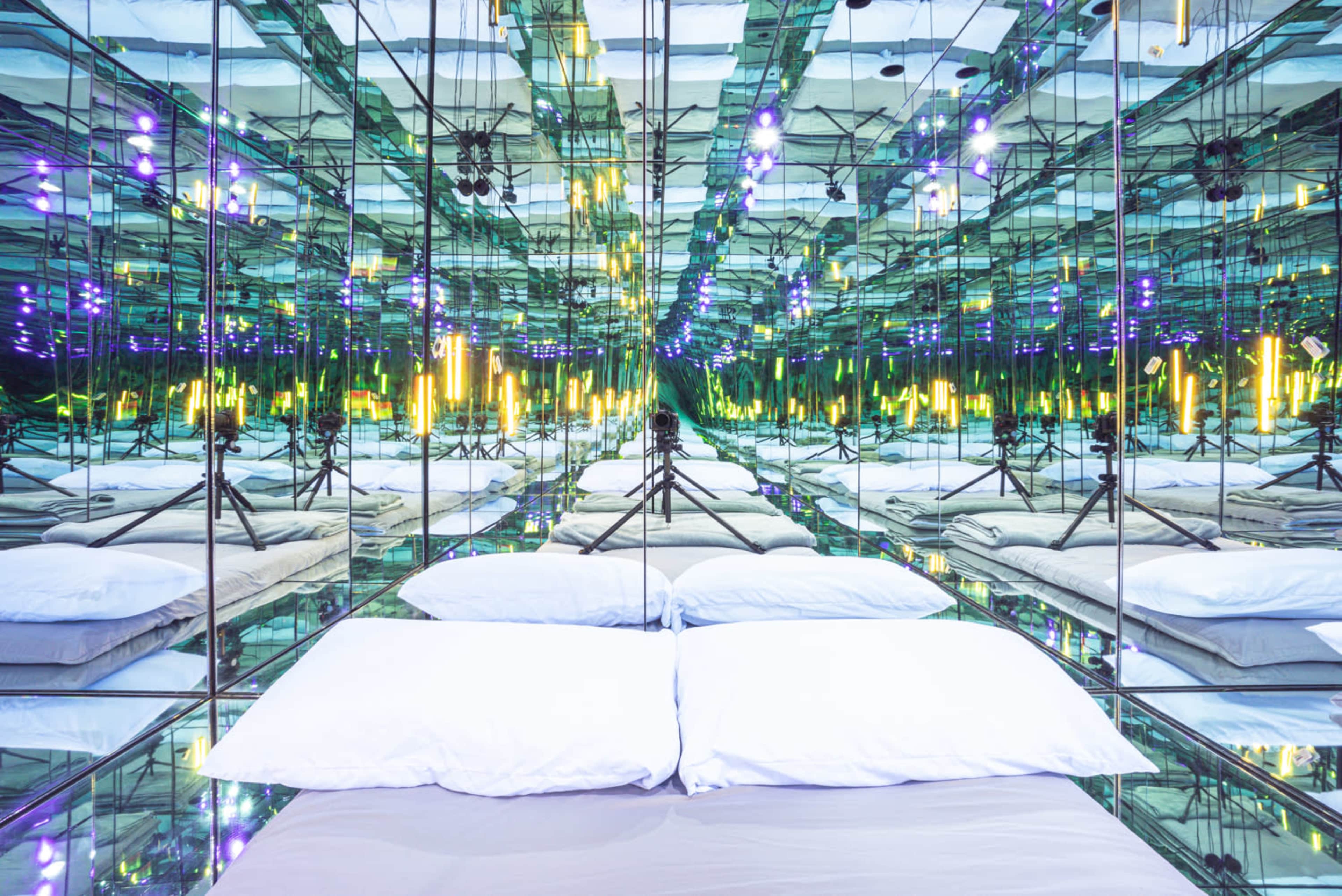 A mirrored room with a bedding area featuring two white pillows and multiple reflections of colorful lights.