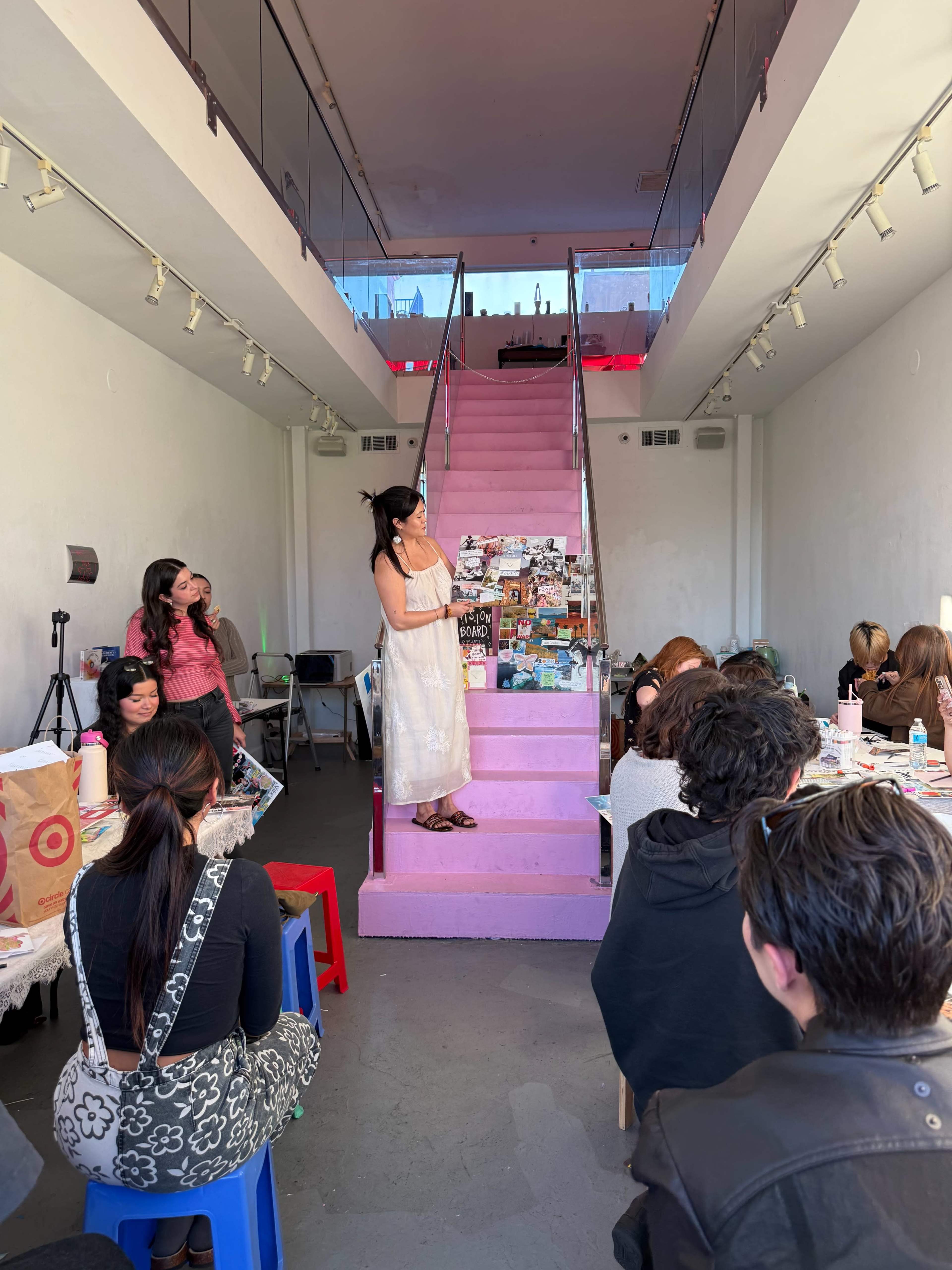 A woman stands on a pink staircase surrounded by an audience seated at tables in a well-lit art space.