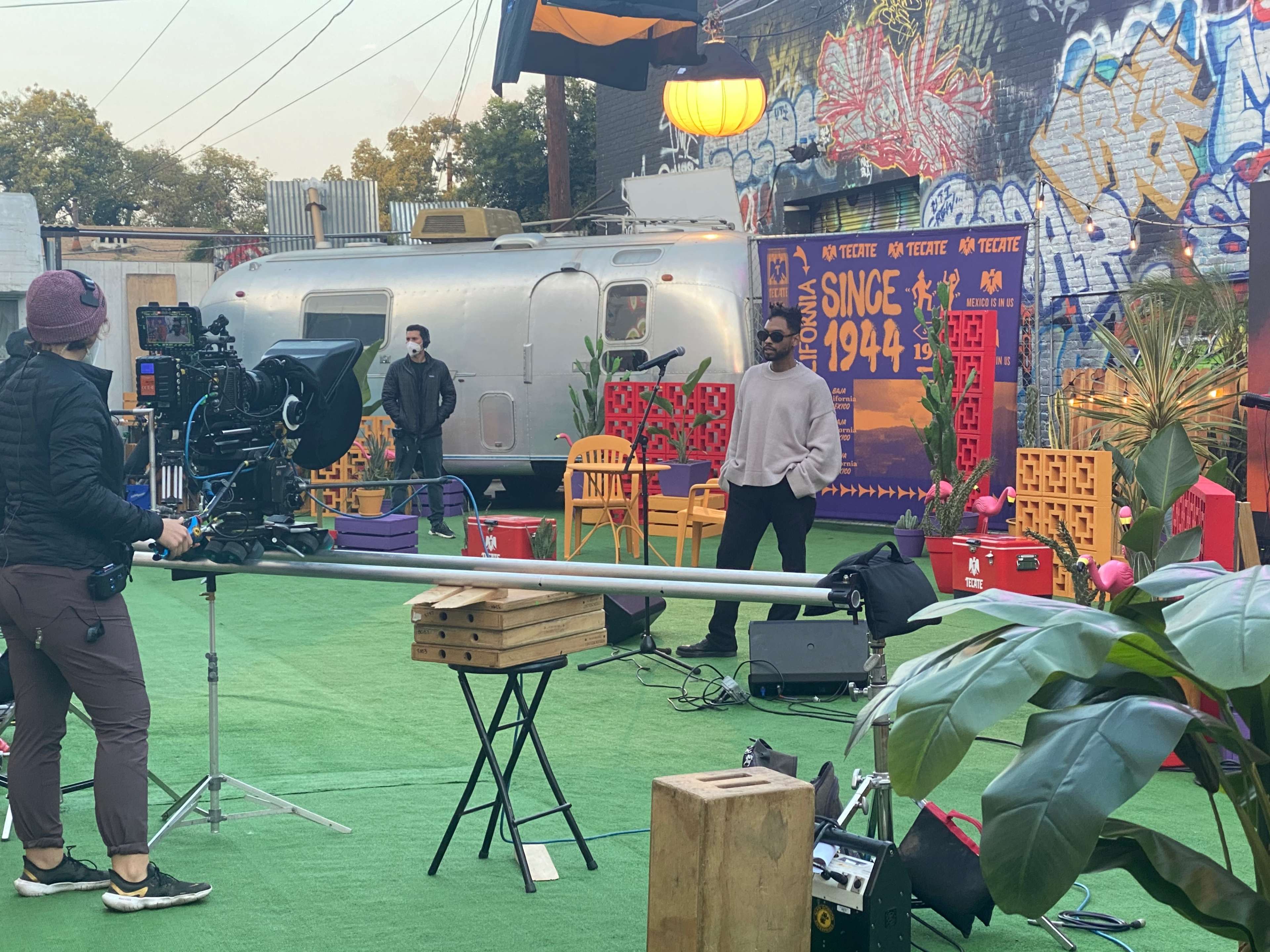 A film crew sets up equipment in an outdoor area featuring colorful decor and a vintage trailer.