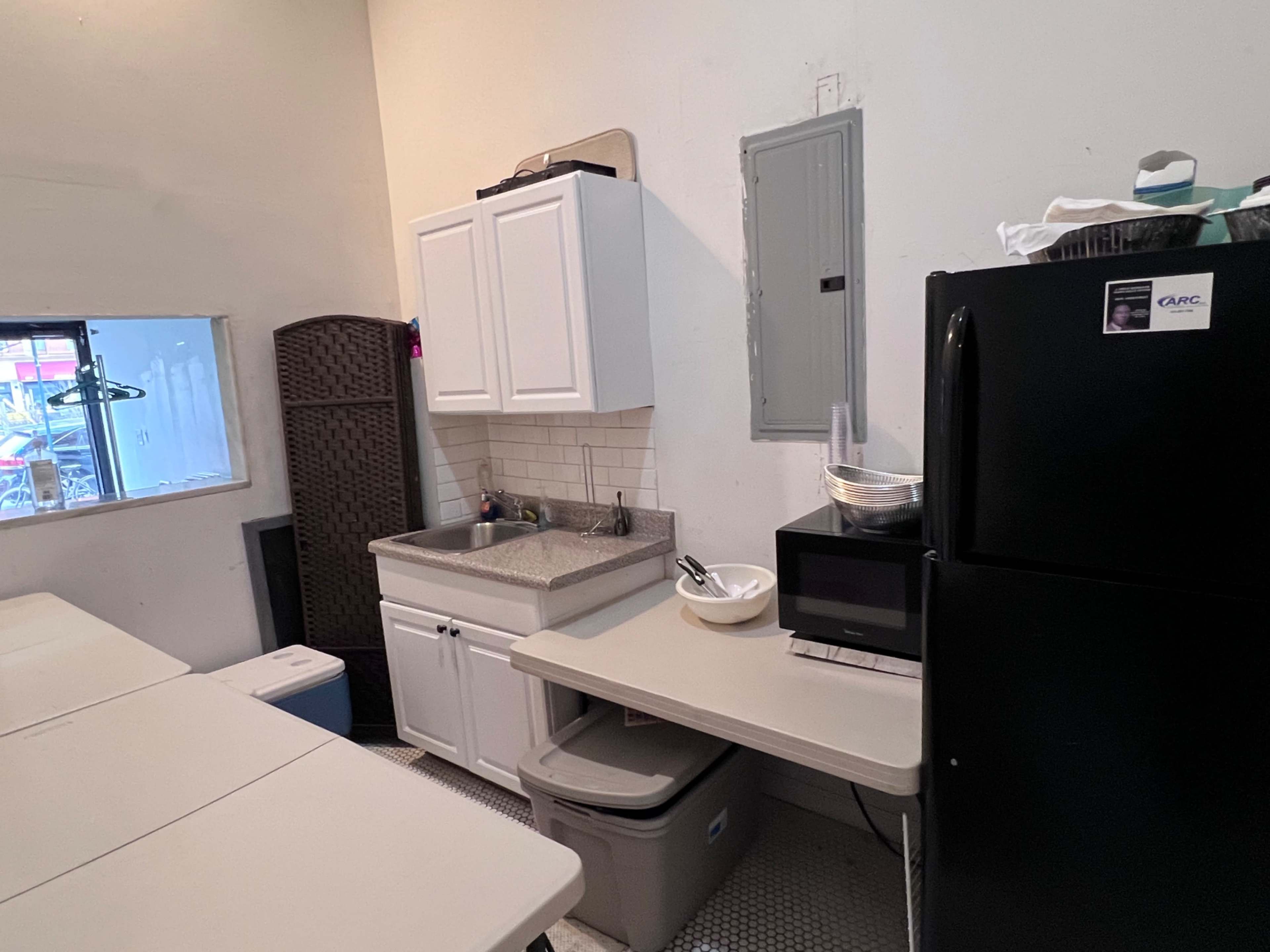 A small kitchen area with a sink, cabinets, a microwave, and a black refrigerator, alongside a table and storage bins.