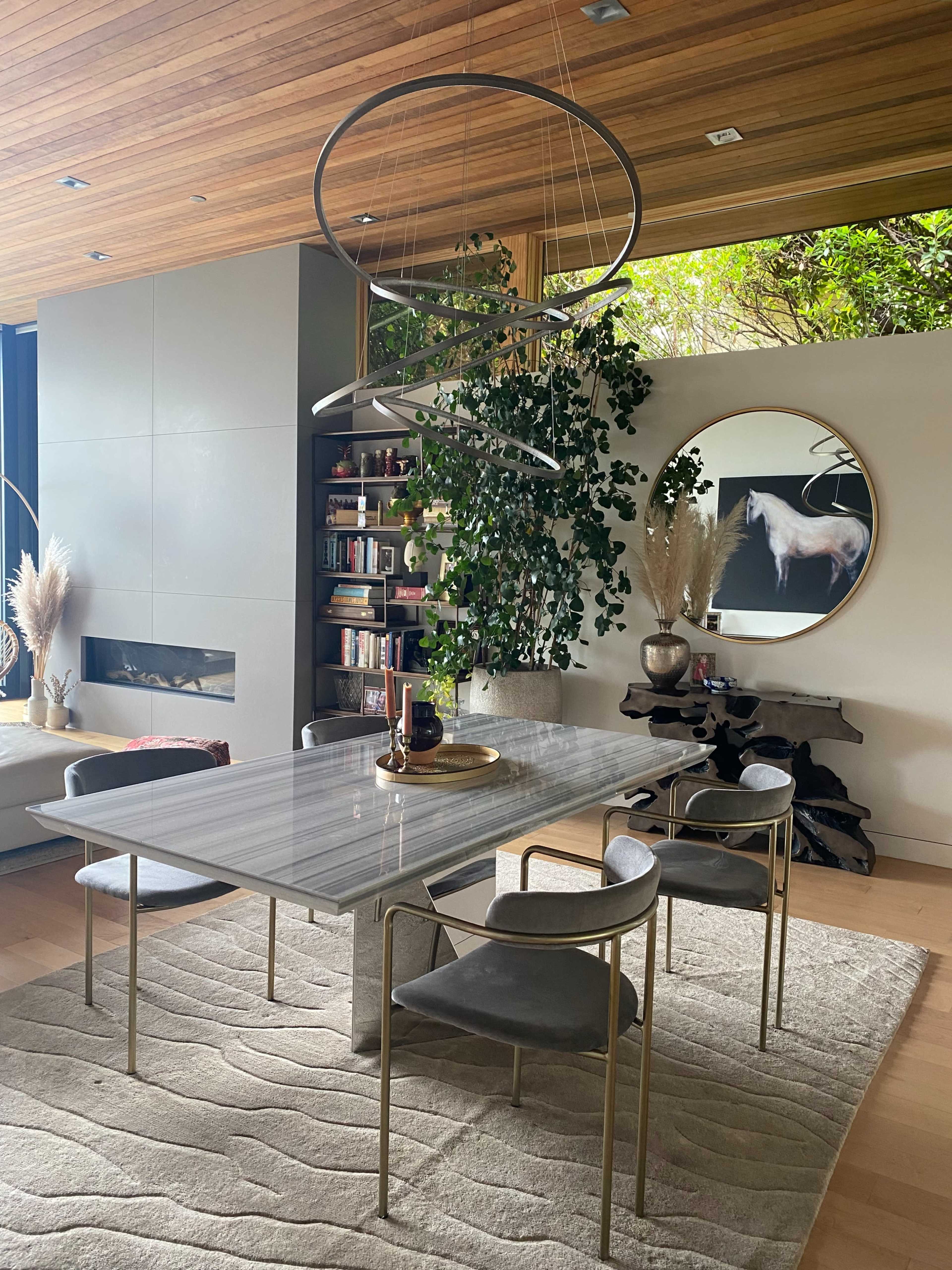 A modern dining room features a marble table surrounded by four chairs, with a large circular mirror and a plant in the background.