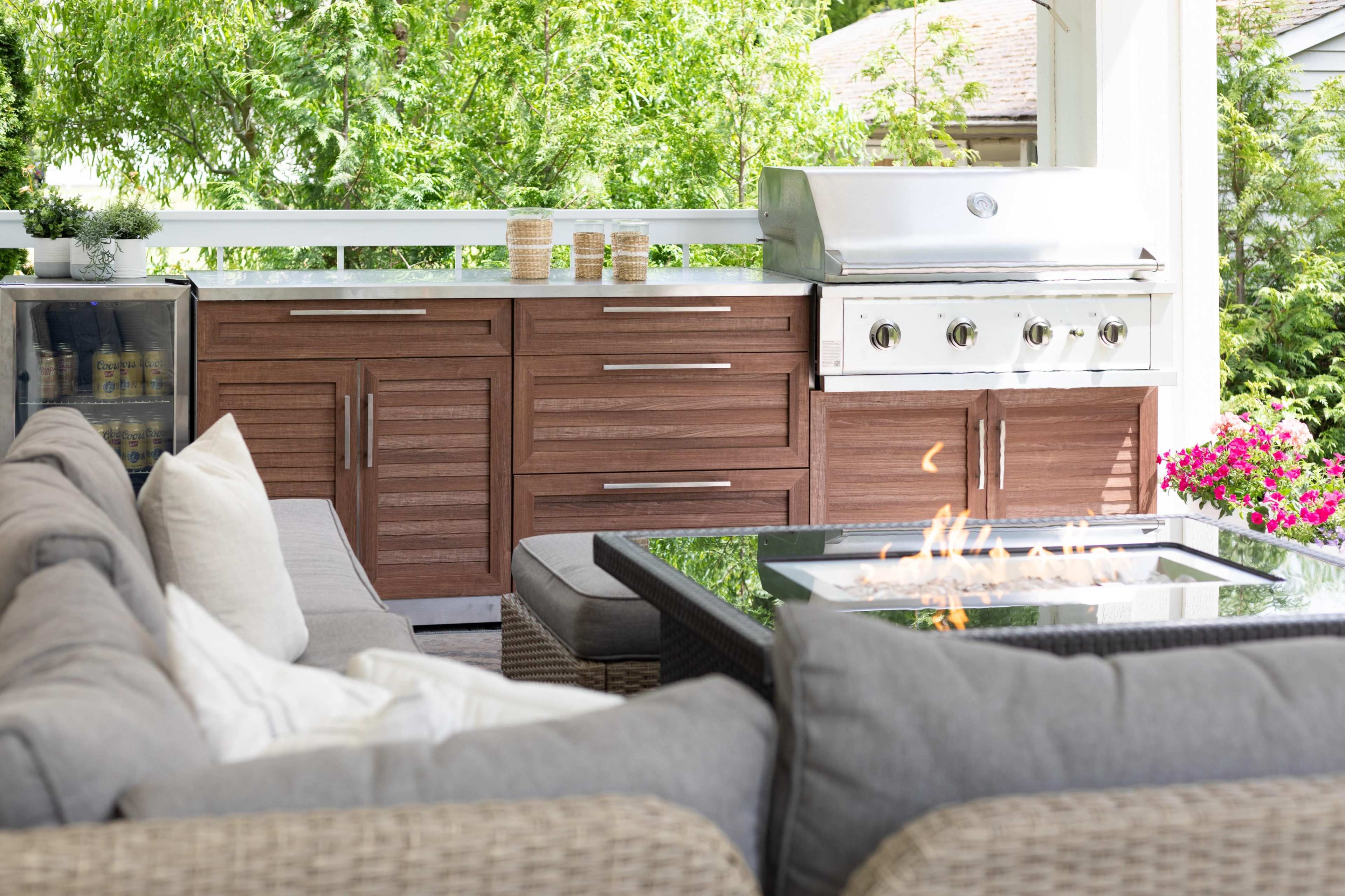 The image shows an outdoor kitchen area featuring a grill, storage cabinets, a fire table, and a comfortable seating arrangement on a patio.