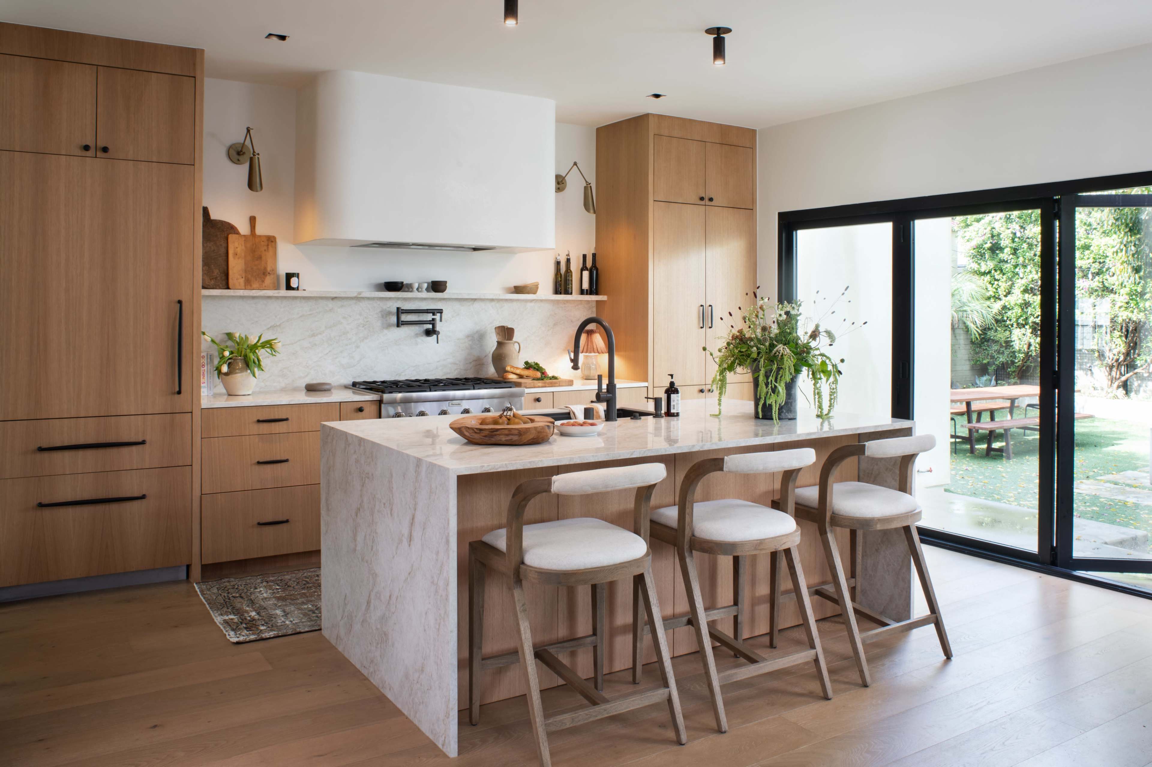 The image shows a modern kitchen featuring wooden cabinetry, a marble island with seating, and large windows overlooking a garden.