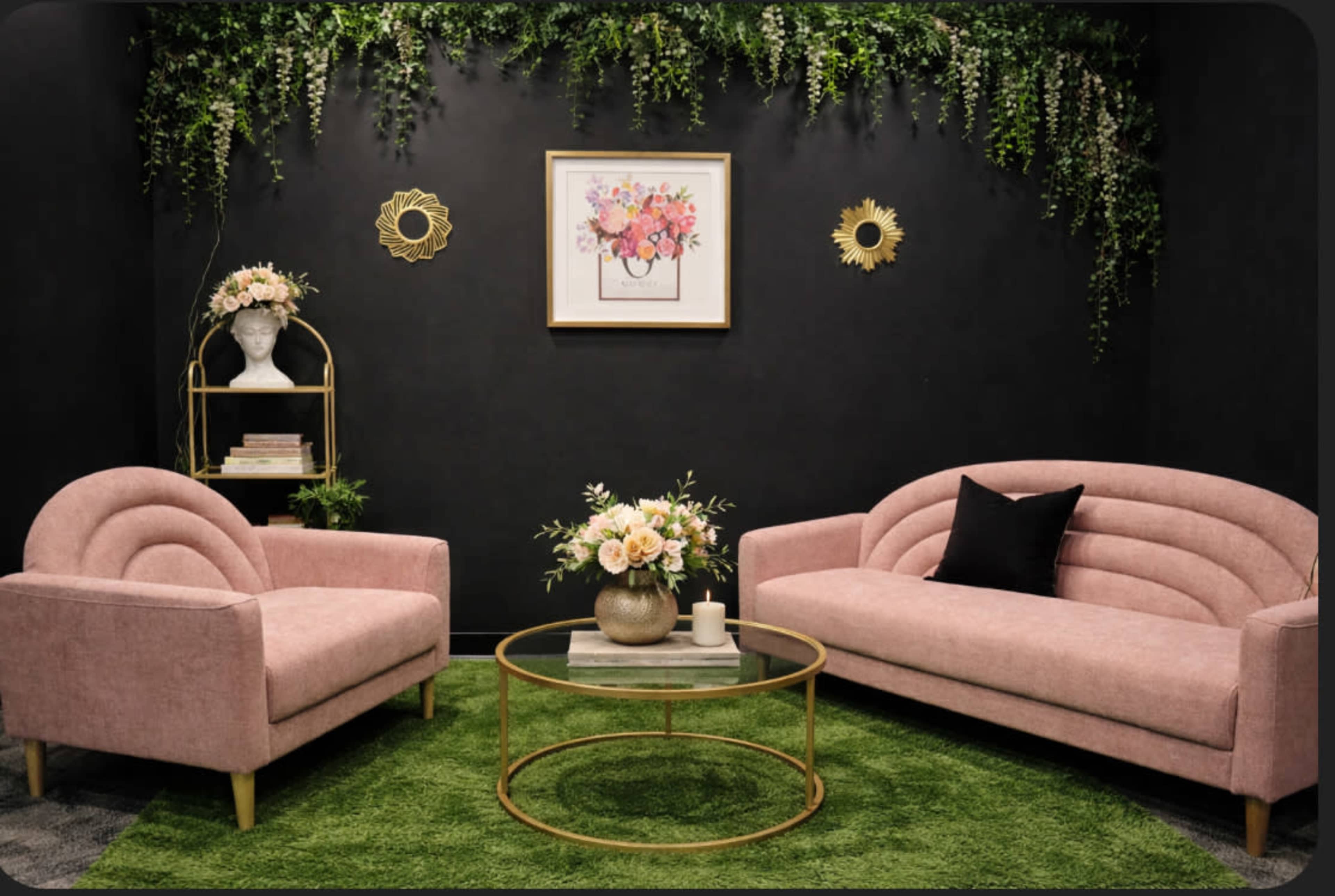 The image shows a stylish interior space featuring two pink sofas, a round glass coffee table, a floral arrangement, and decorative wall art against a dark background with greenery.