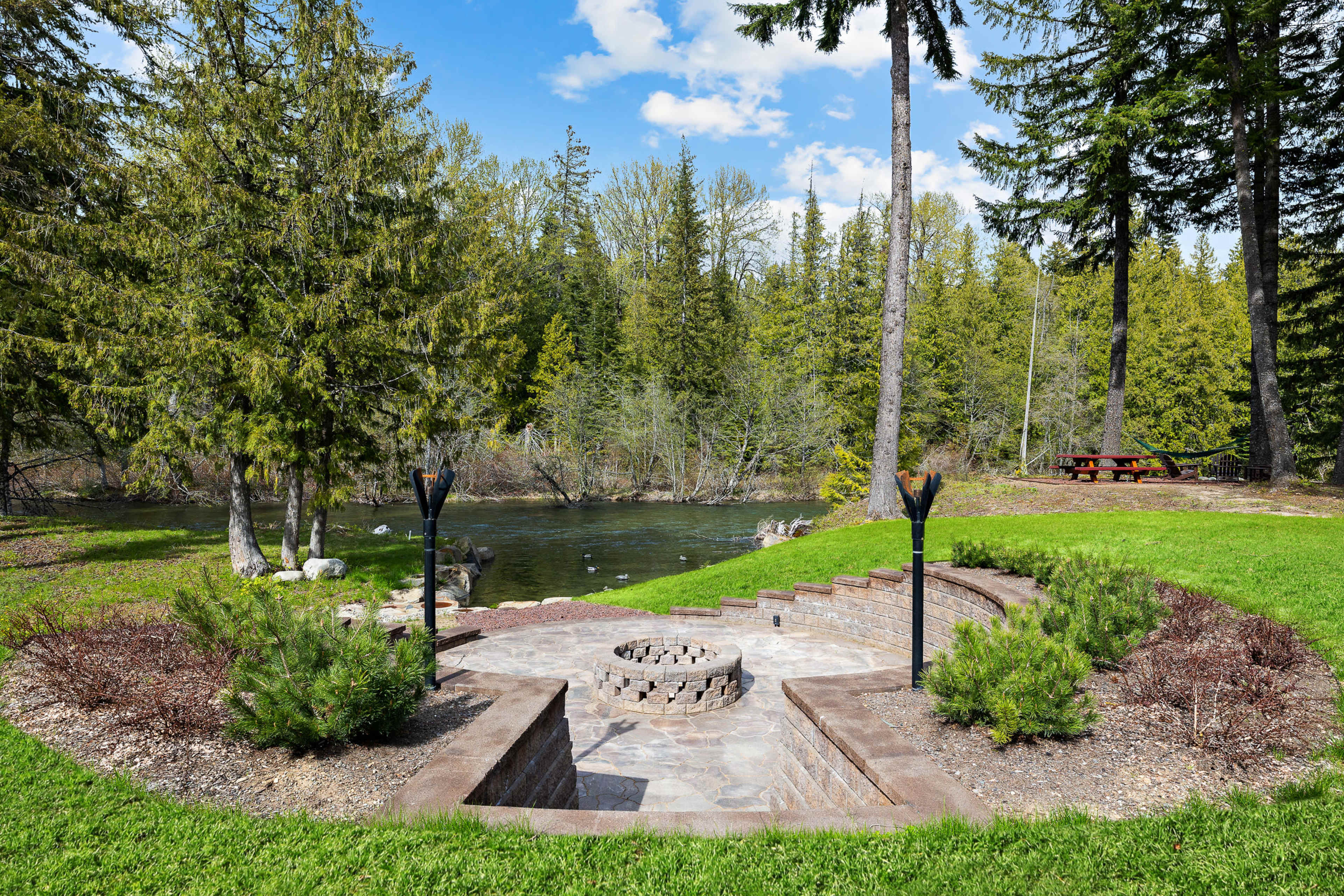 A stone patio with a circular fire pit overlooks a calm pond surrounded by trees and a picnic area.