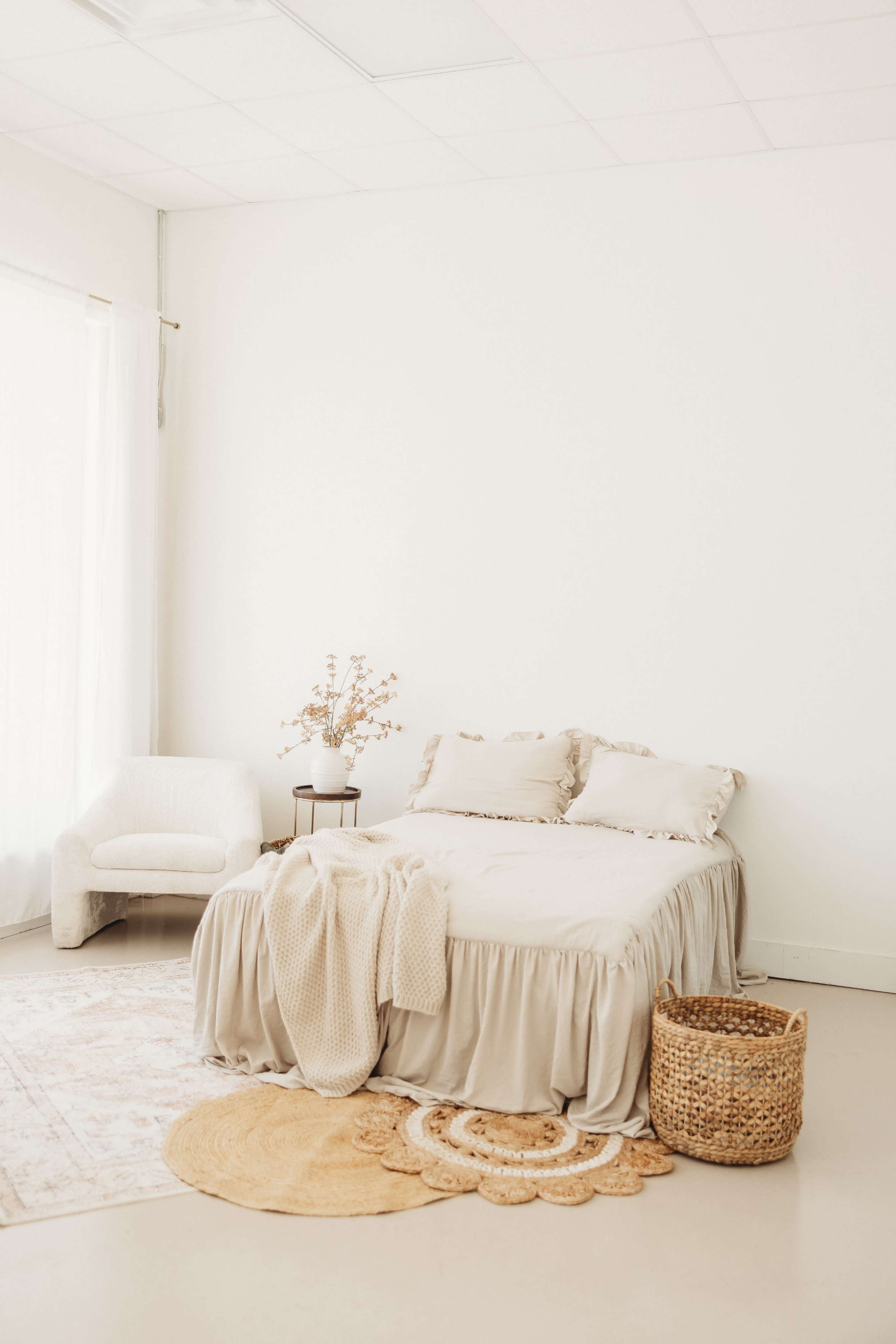 The image shows a minimalist bedroom featuring a beige bedspread, a small round side table with a vase of dried flowers, and a woven basket on a circular rug.