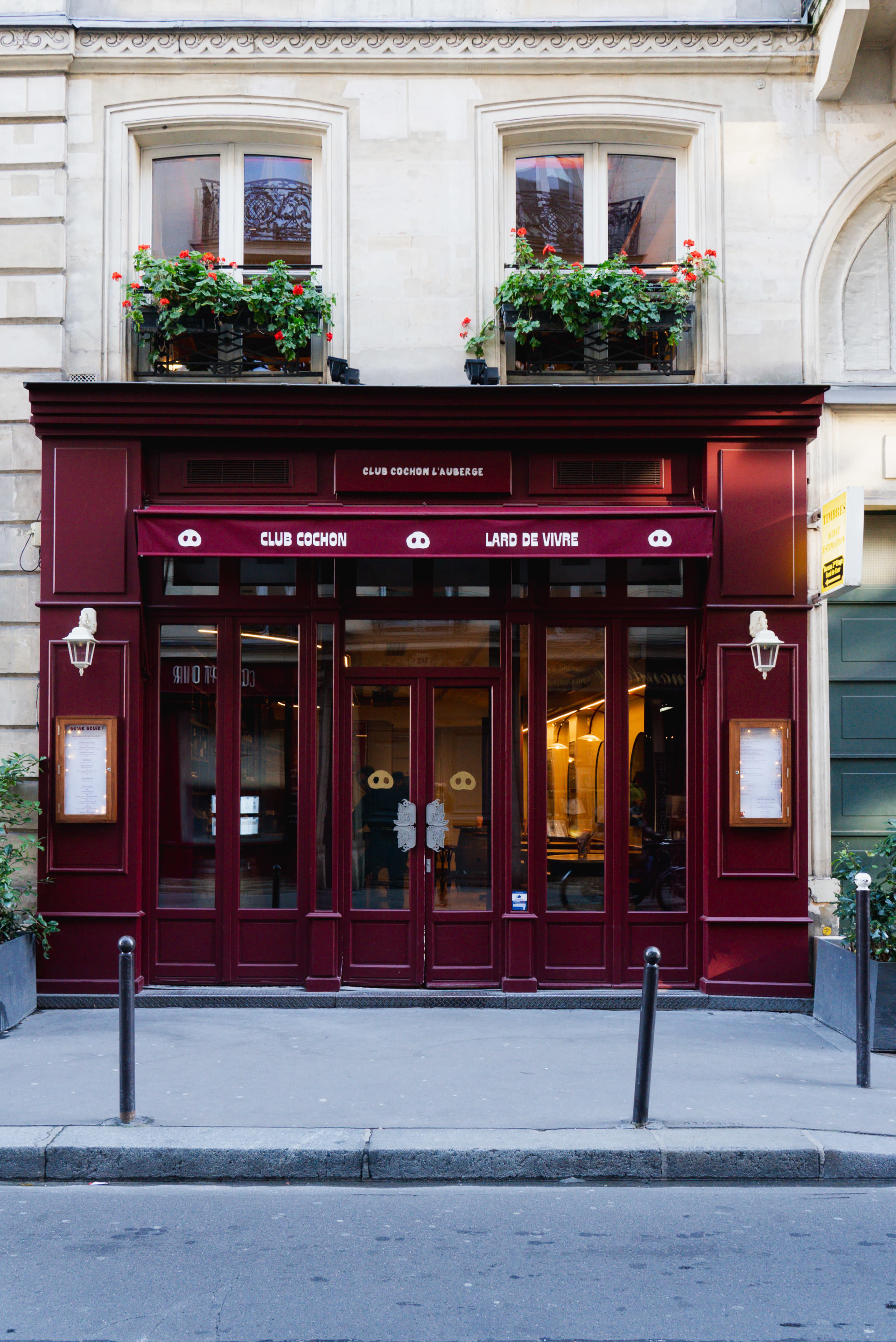 The image shows a red storefront with a canopy that reads "Club Cochon" and "L'Art de Vivre," featuring decorative flower planters above the entrance.