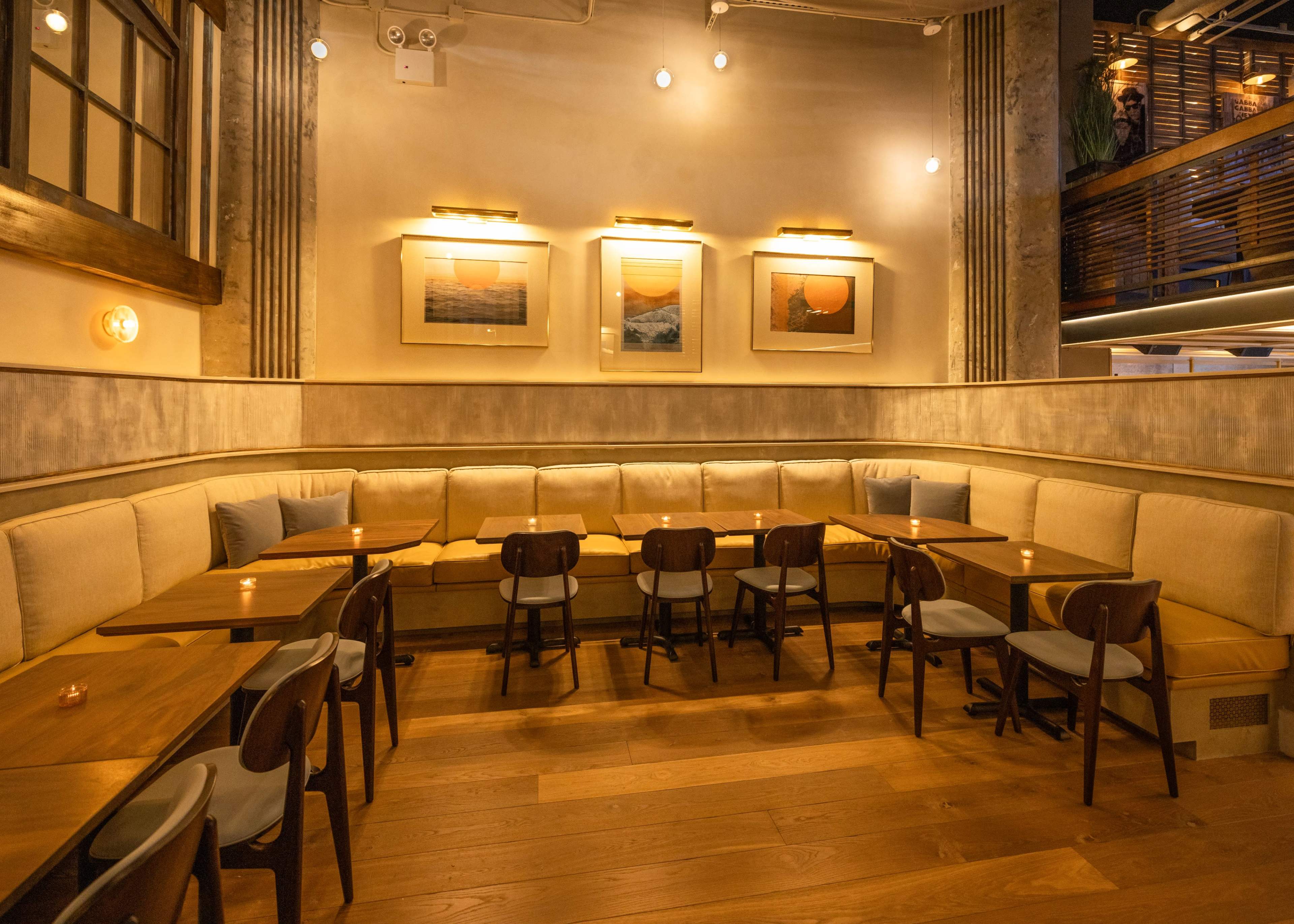 A cozy restaurant interior with wooden tables and chairs arranged around a padded bench along the wall, featuring framed artwork and soft lighting.